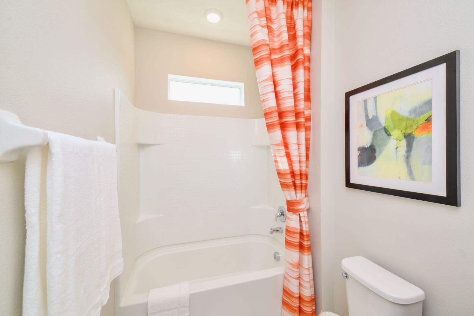 Compact bathroom with a white shower/tub combo, a vibrant orange and white striped shower curtain, and a toilet. A small horizontal window is visible above the shower.