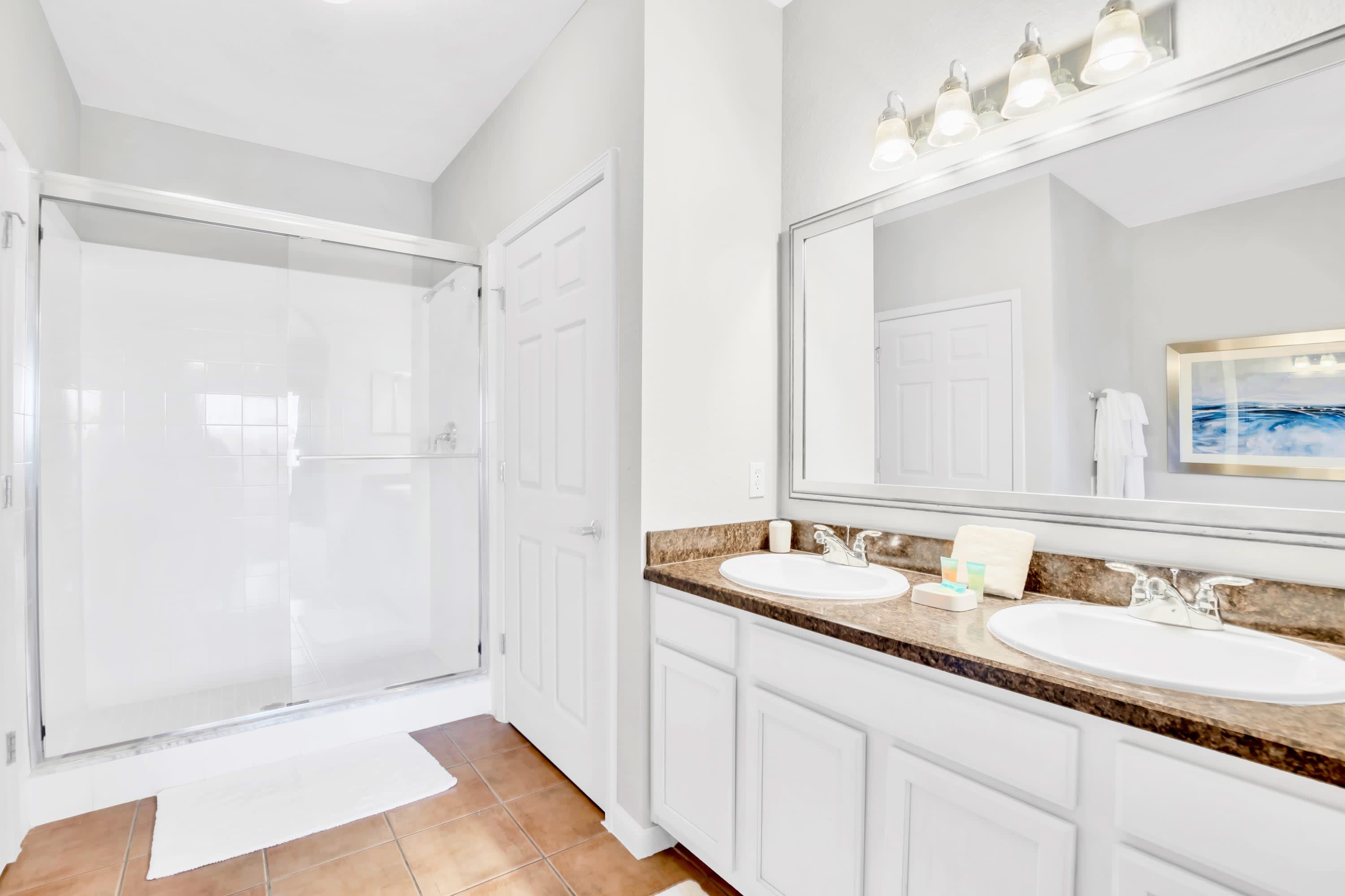 Modern bathroom with a double vanity featuring white sinks set into speckled brown countertops, a large mirror, and a glass-enclosed walk-in shower. The floor is tiled in light brown.