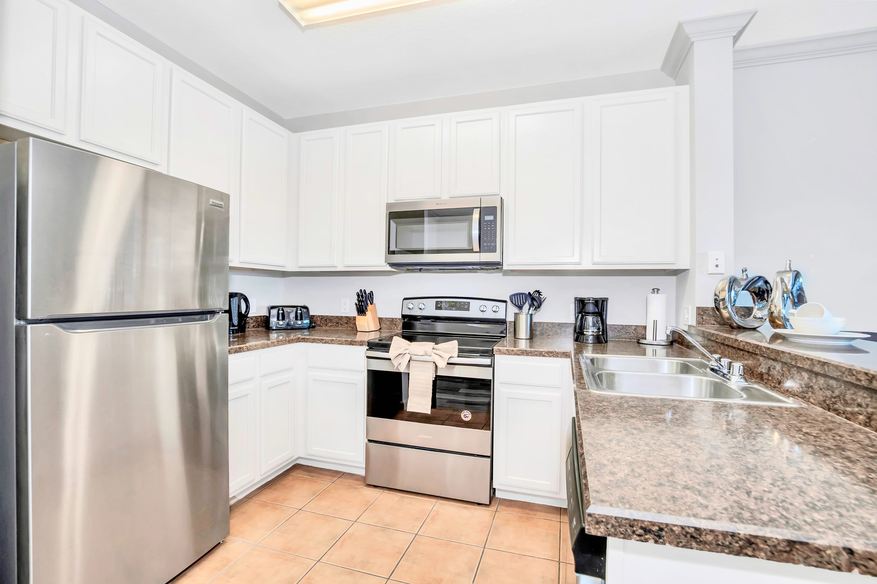 Modern kitchen with white upper and lower cabinets, stainless steel appliances including a refrigerator, oven, microwave, and dishwasher, and speckled brown granite countertops. The floor is covered in light brown square tiles.