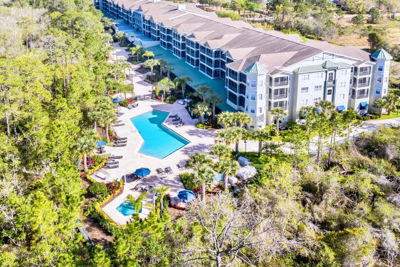 Aerial view of a large multi-story condominium complex with a long, winding outdoor swimming pool and hot tub, surrounded by lush green trees and tropical landscaping.