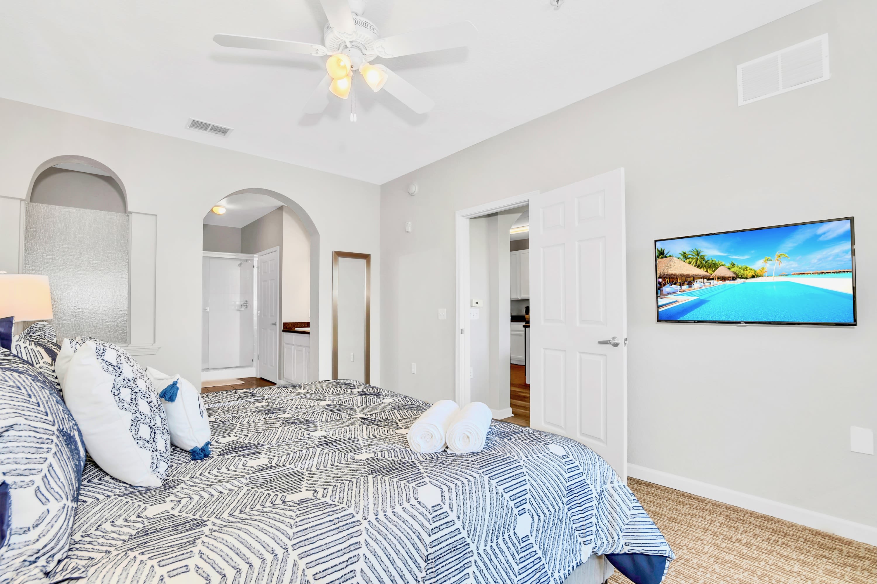 Bright bedroom with a large bed featuring a blue and white geometric patterned comforter, white pillows, and a wall-mounted flat-screen TV displaying a beach scene. An arched doorway leads to an en-suite bathroom.