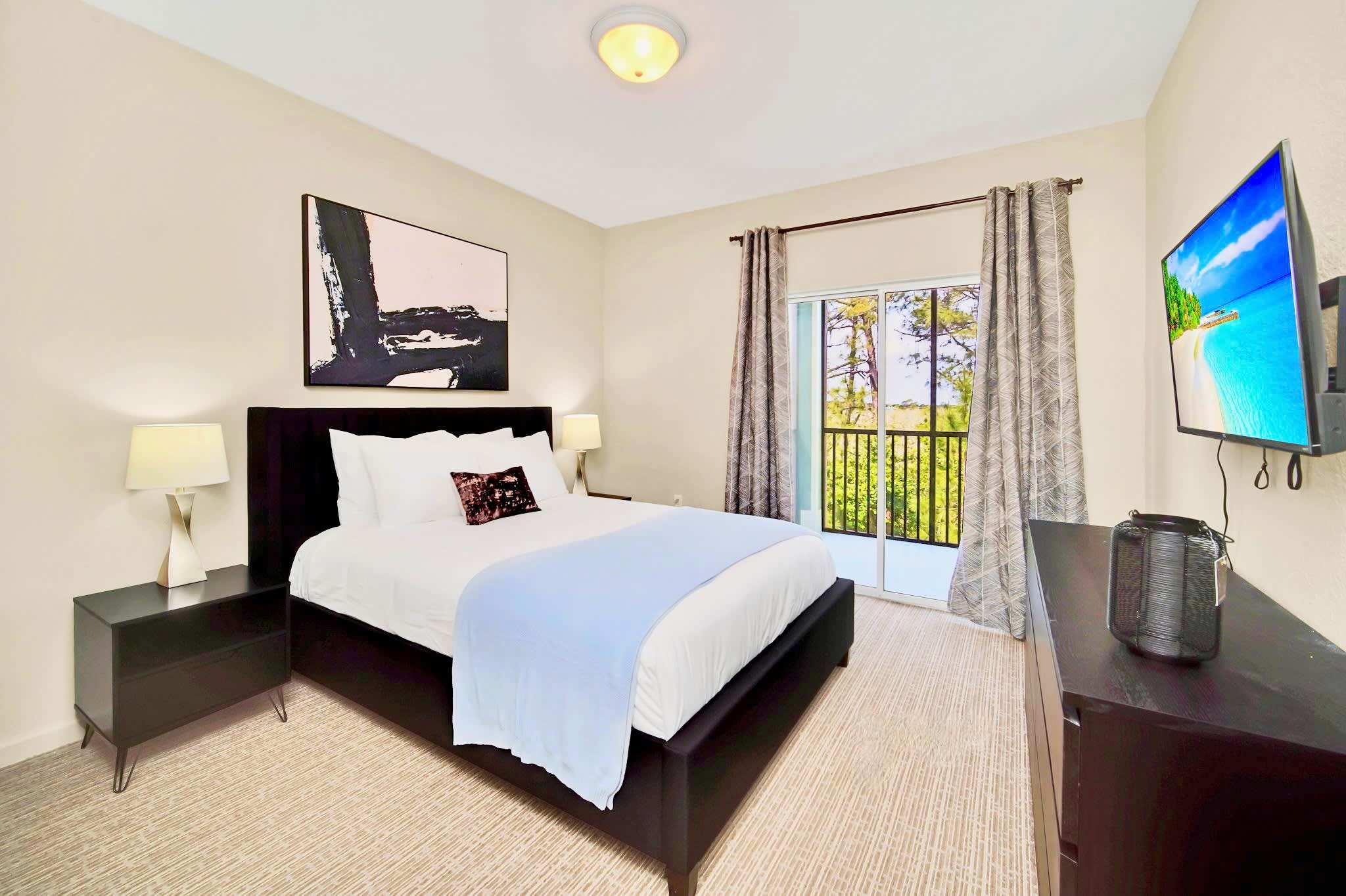 A cozy bedroom with a black upholstered bed, white bedding, and a light blue throw, flanked by black nightstands with lamps. A large abstract black and white artwork hangs above the bed, and a balcony with a view of trees is accessible through glass doors.