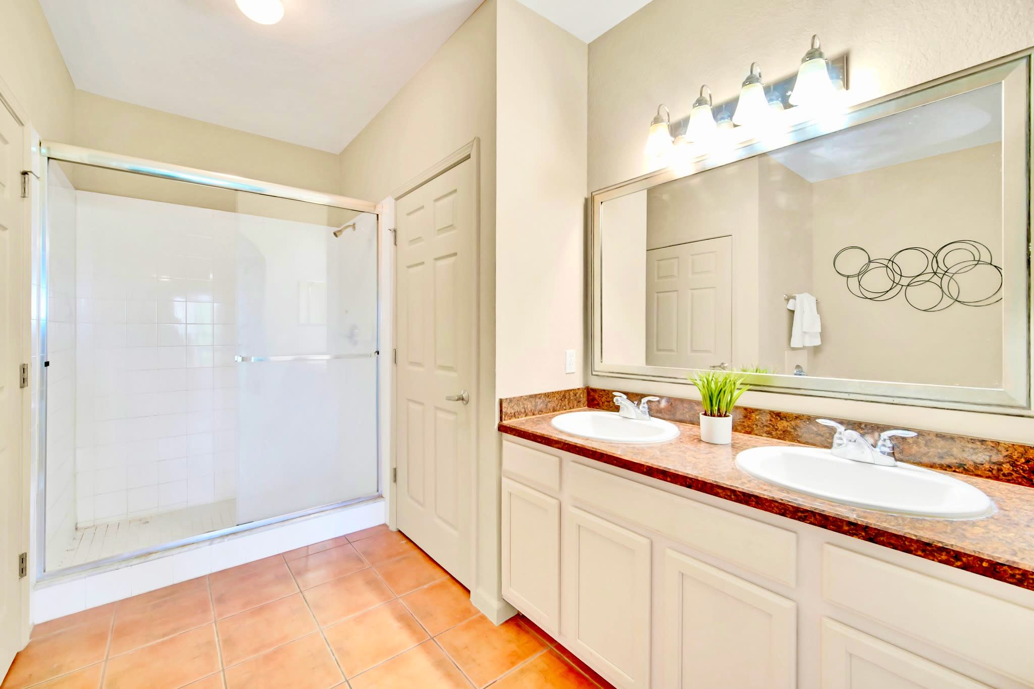 A bright bathroom featuring a double vanity with a brown speckled countertop, white sinks, and a large mirror with a light fixture above. A glass-enclosed shower with white tiles is visible on the left.