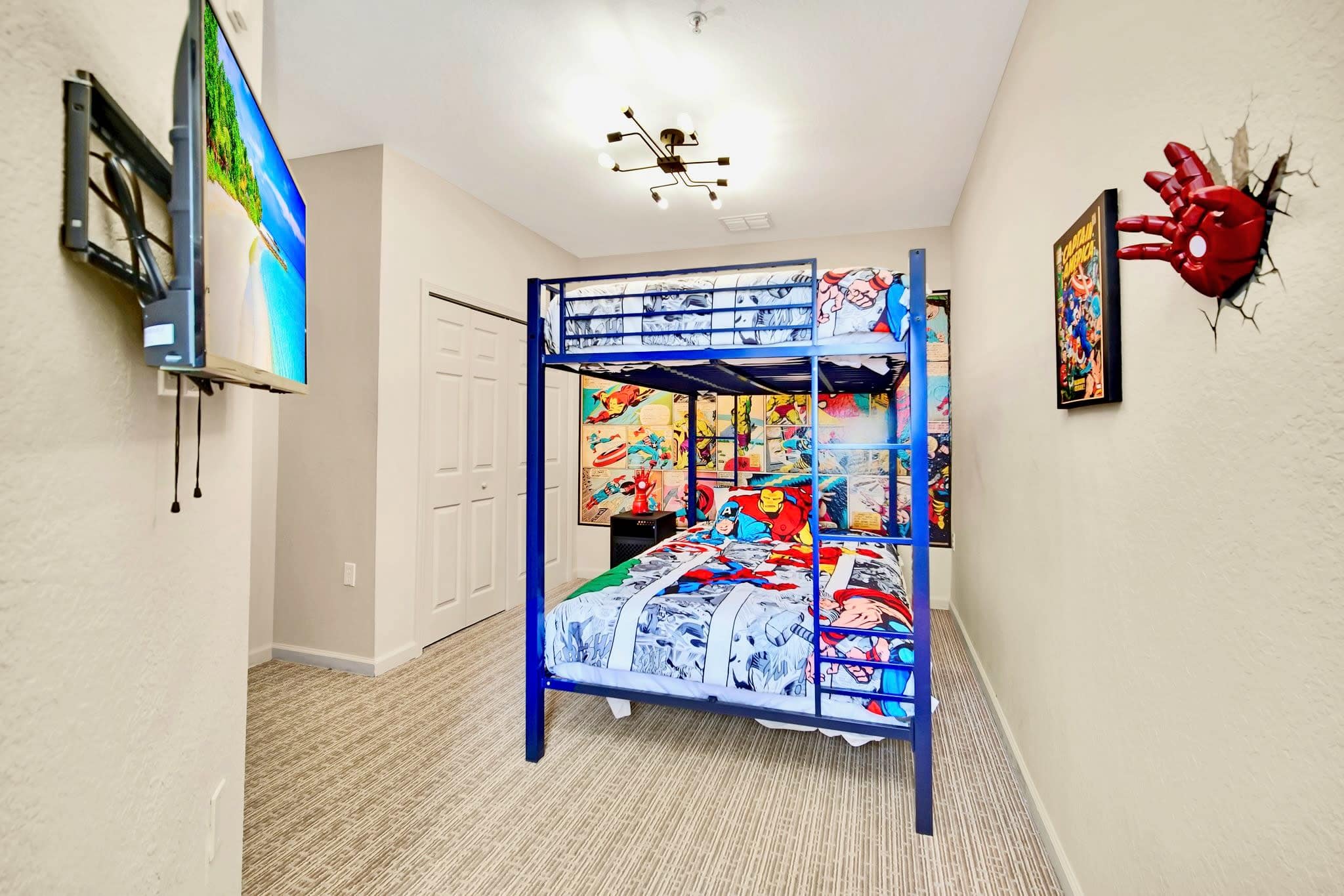 A vibrant kids' bedroom featuring a blue metal bunk bed with superhero-themed bedding. A wall-mounted TV is on the left, and a 3D Iron Man hand light fixture is on the right wall, adding a playful touch.