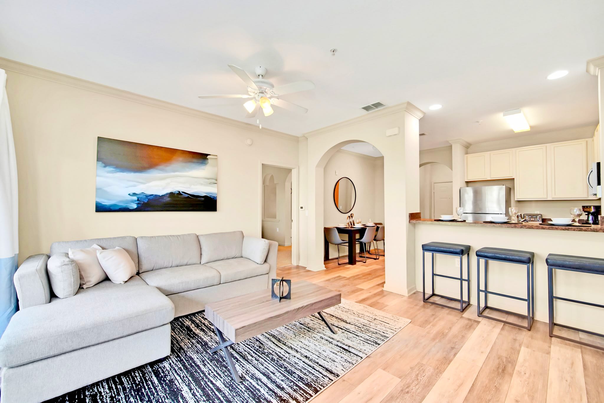 An open-concept living space with a light grey sectional sofa, a wooden coffee table, and an abstract painting, flowing into a kitchen with a breakfast bar. Hardwood floors extend through the living and dining areas.