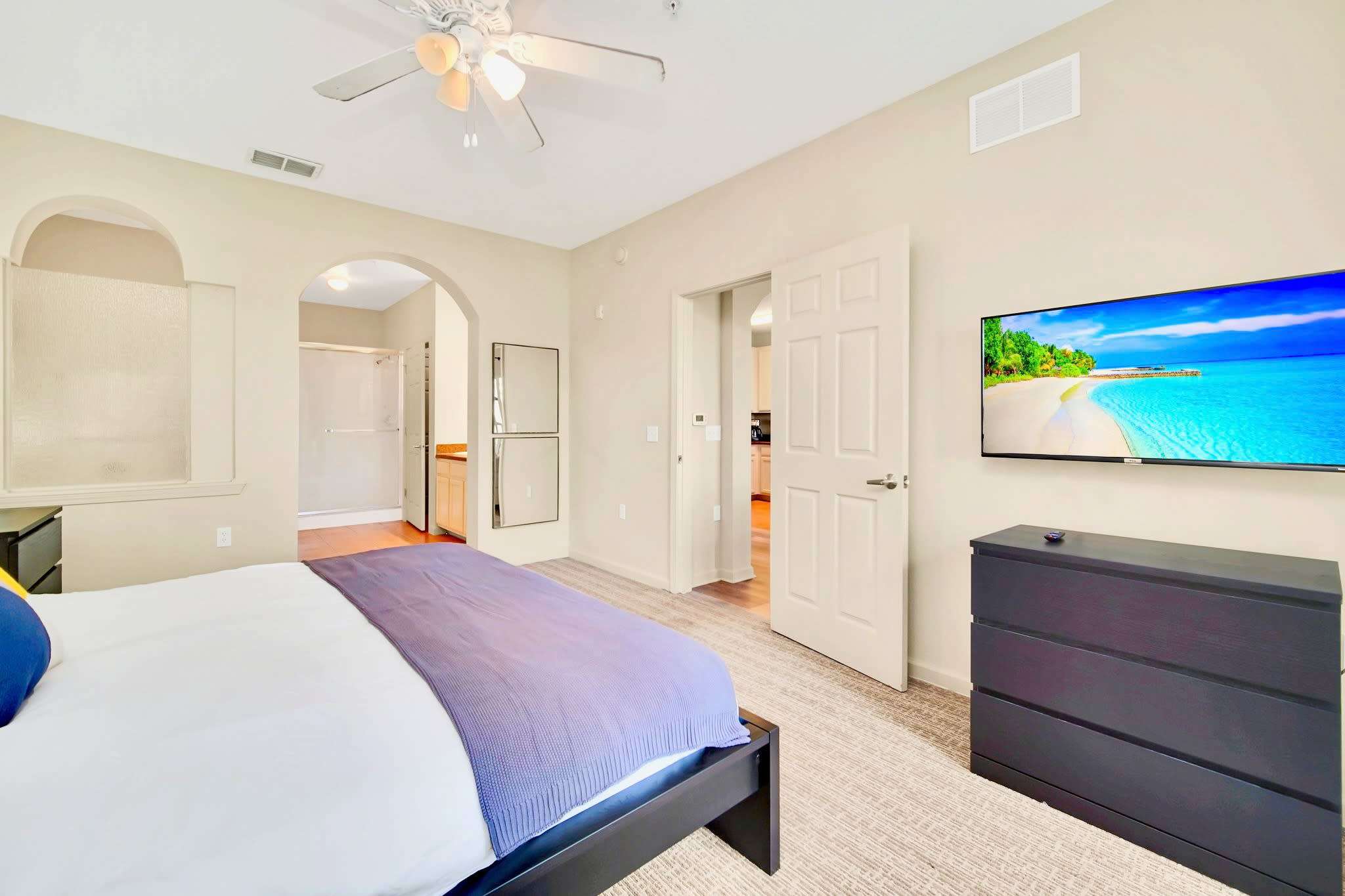The master bedroom features a dark wood bed frame, a wall-mounted TV, and a dark dresser, with an arched opening leading to the en-suite bathroom. Carpeted floors and light-colored walls create a calm atmosphere.