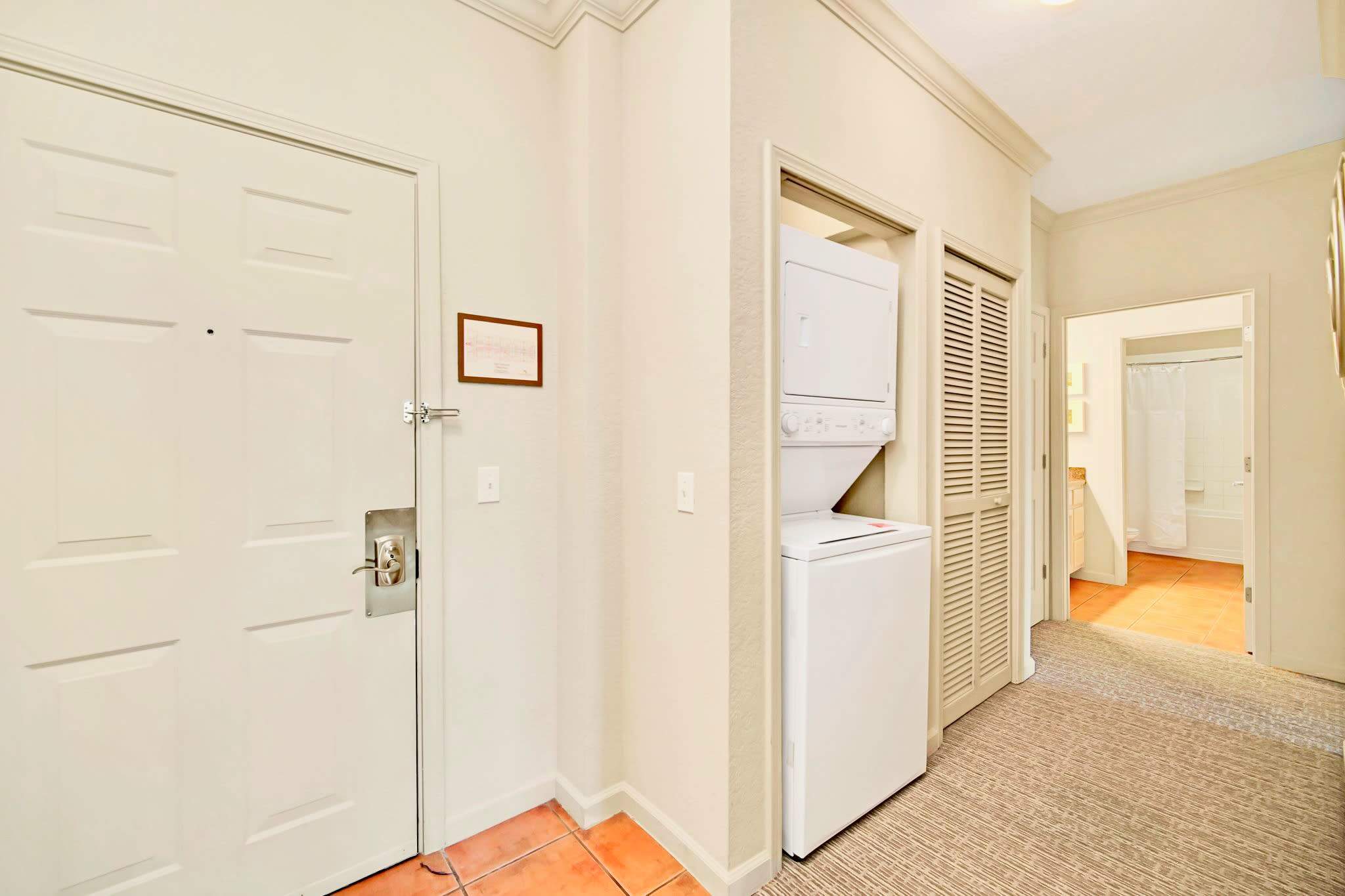 A functional entryway or hallway featuring a stacked white washer and dryer unit in a closet next to the main entrance door. The floor transitions from tile to carpet, and a closed louvered door is also visible.