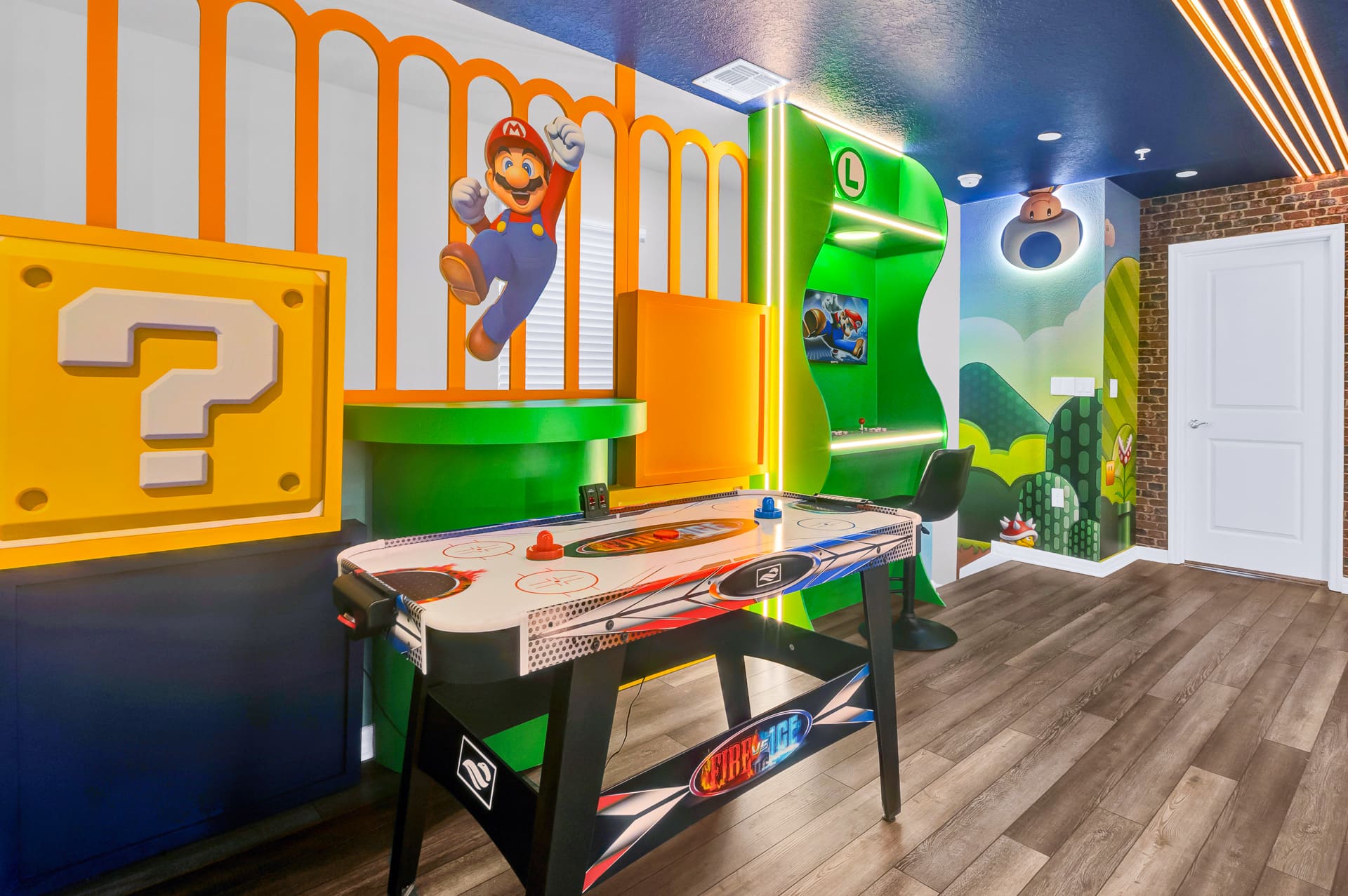 A colorful Mario-themed game room showcases an air hockey table in the foreground, a large green arcade cabinet, and vibrant wall murals with Mario characters and question blocks. The room features dark wood-look flooring and bright LED strip lighting on the ceiling.