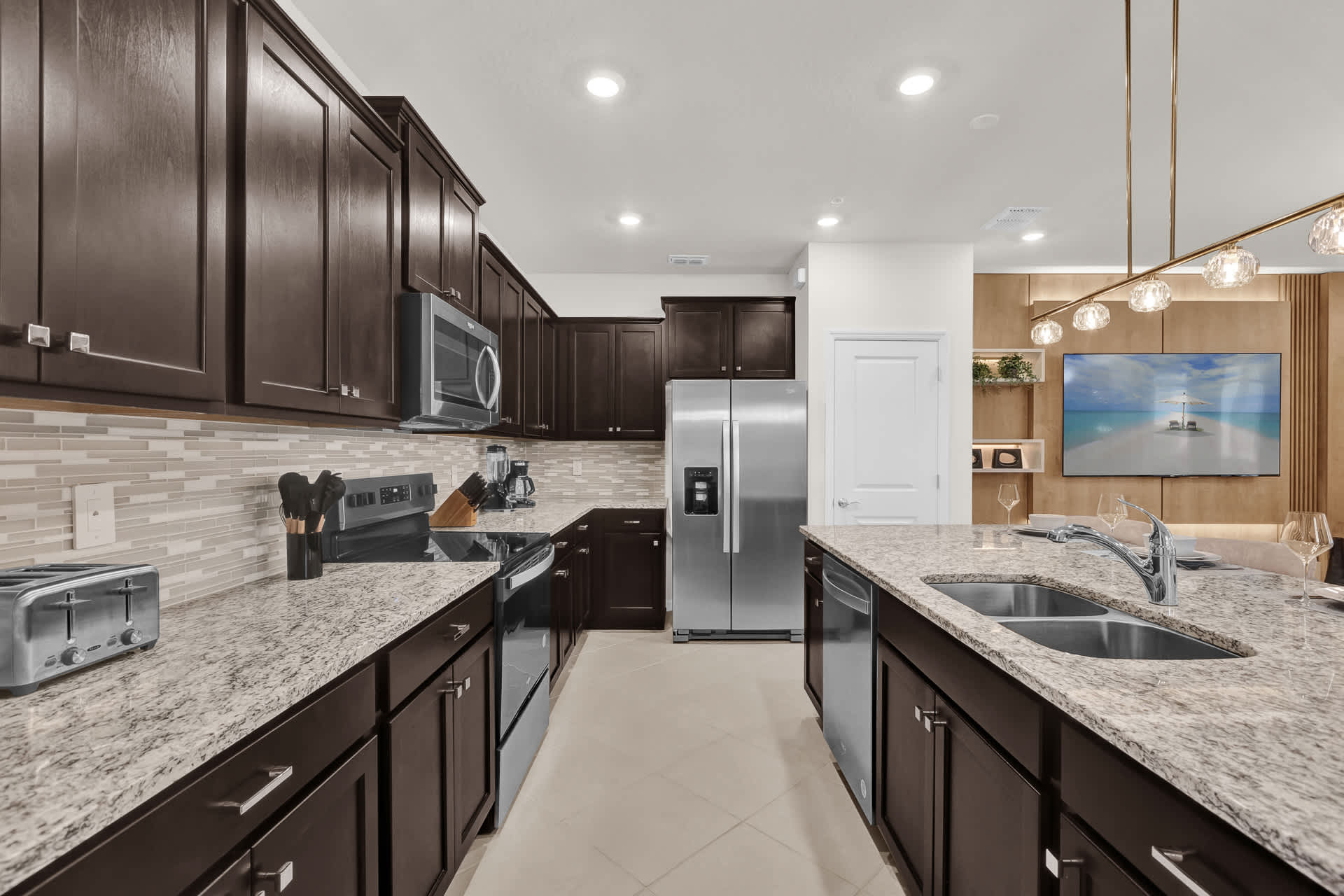 A well-equipped kitchen showcases dark wood cabinetry, light granite countertops, and a subway tile backsplash, with a stainless steel refrigerator and range. The large kitchen island features a double sink and ample prep space, illuminated by recessed lighting.