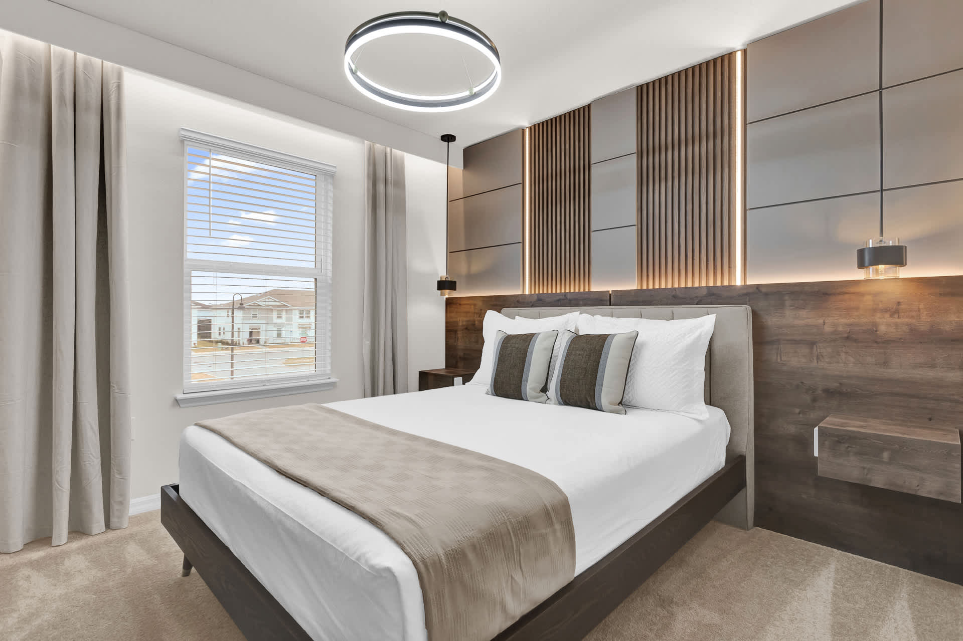 Modern bedroom with a queen bed, a decorative wood and gray paneled accent wall with integrated lighting, and a large window with blinds and sheer curtains. A circular LED chandelier hangs from the ceiling.