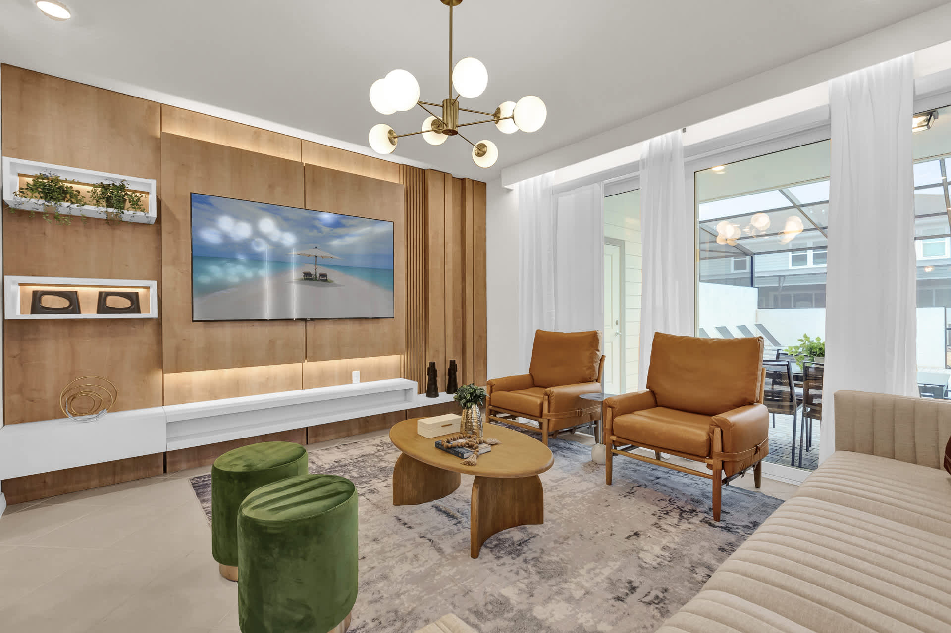 A modern living room features a large wall-mounted TV on a light wood paneled accent wall with integrated lighting and a white media console. Two tan leather armchairs, two green velvet ottomans, and a light-colored sofa surround a wooden coffee table on a patterned rug.