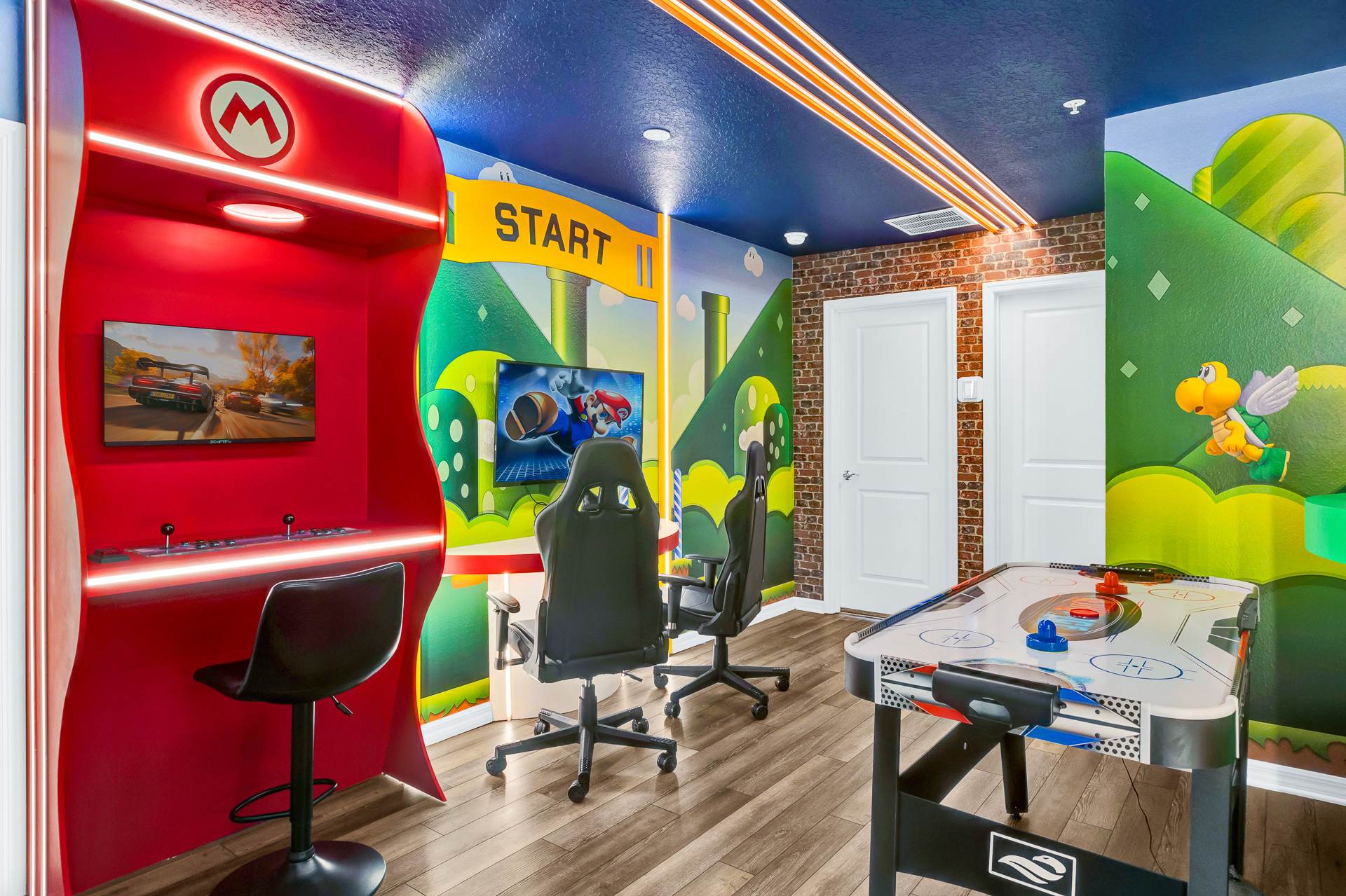 A vibrant Mario-themed game room features a red arcade cabinet with a racing game, two gaming chairs in front of a TV displaying Mario Kart, and an air hockey table. The walls are decorated with Super Mario Bros. landscapes and a brick pattern.