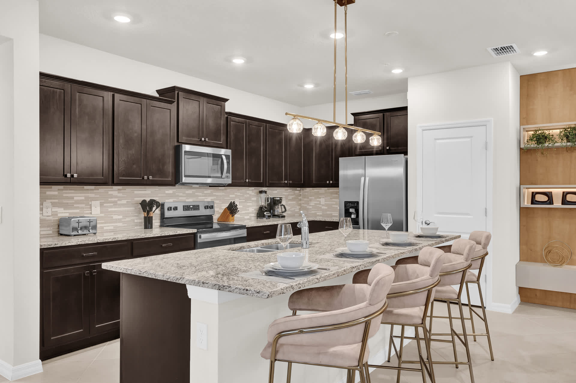 A modern kitchen features dark wood shaker-style cabinets, light granite countertops, and a beige subway tile backsplash. Stainless steel appliances include a refrigerator, microwave, range, and dishwasher, with a large island providing additional counter space and seating.