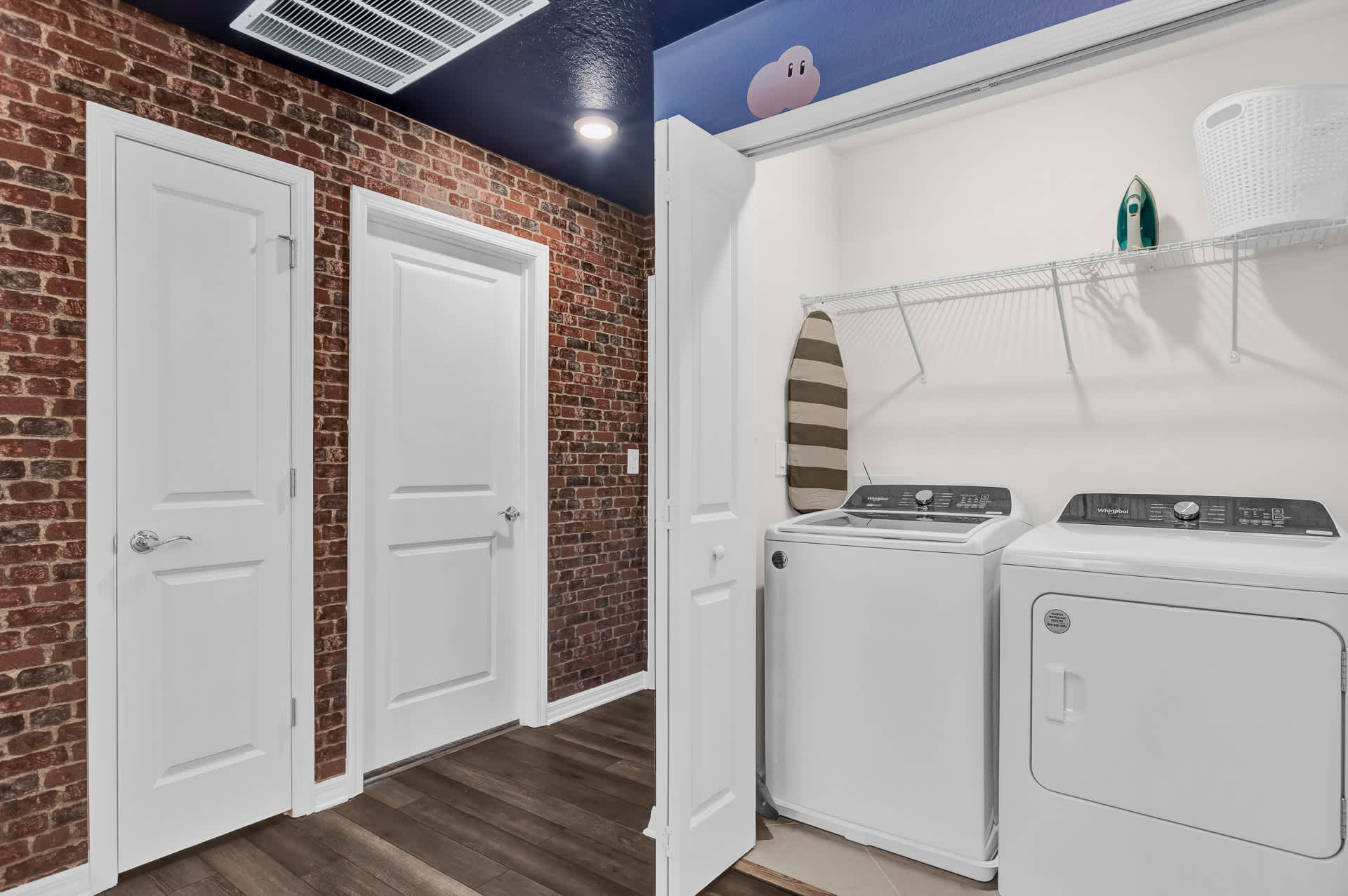 Compact laundry area with a white top-loading washing machine and a matching dryer, an ironing board, and shelving. The adjacent hallway features a rustic exposed brick wall and dark wood flooring.