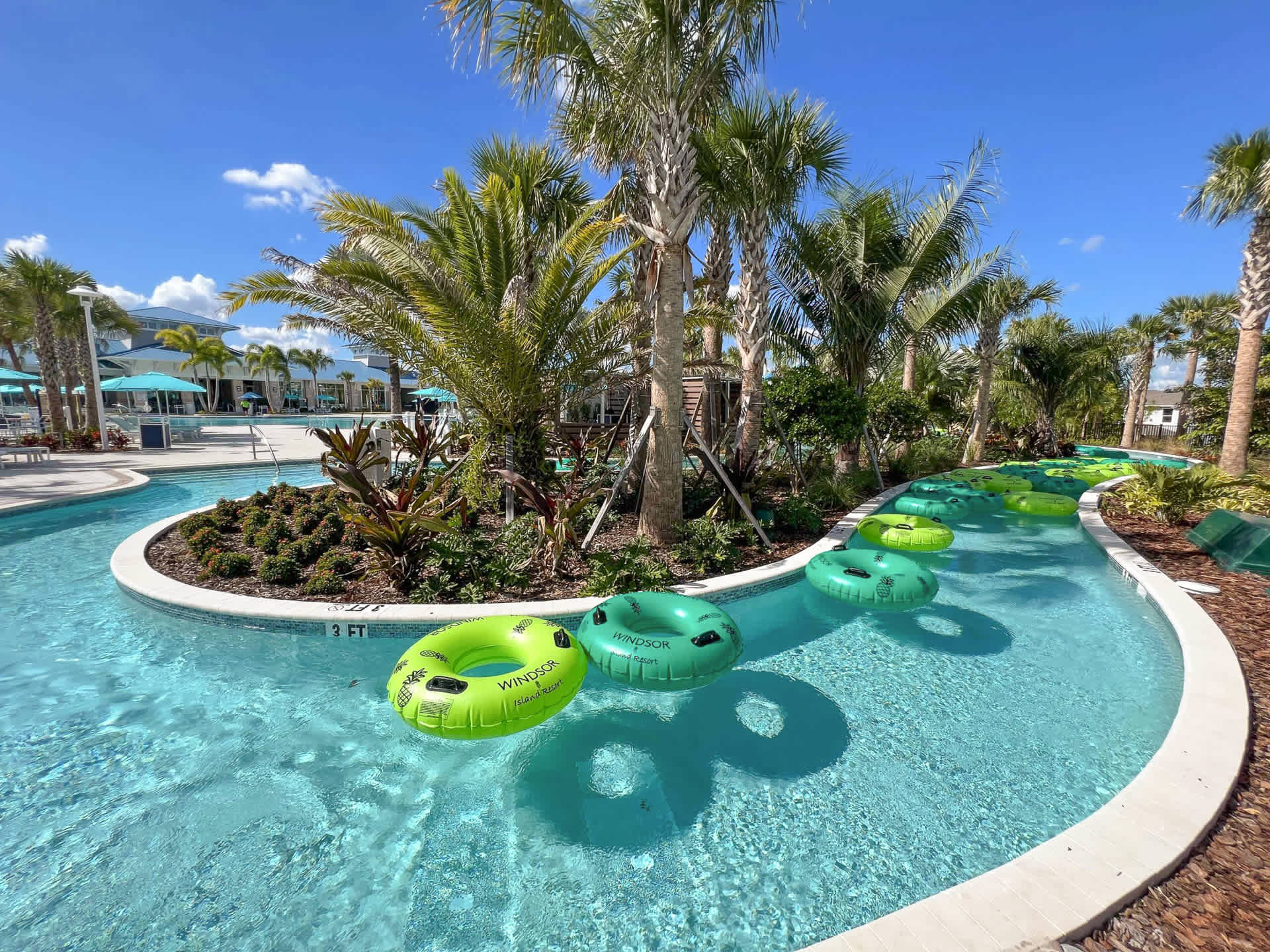 Serene view of a lazy river with several green inner tubes floating in the clear blue water, surrounded by lush tropical plants and palm trees under a sunny sky. The main pool area is visible in the distance.