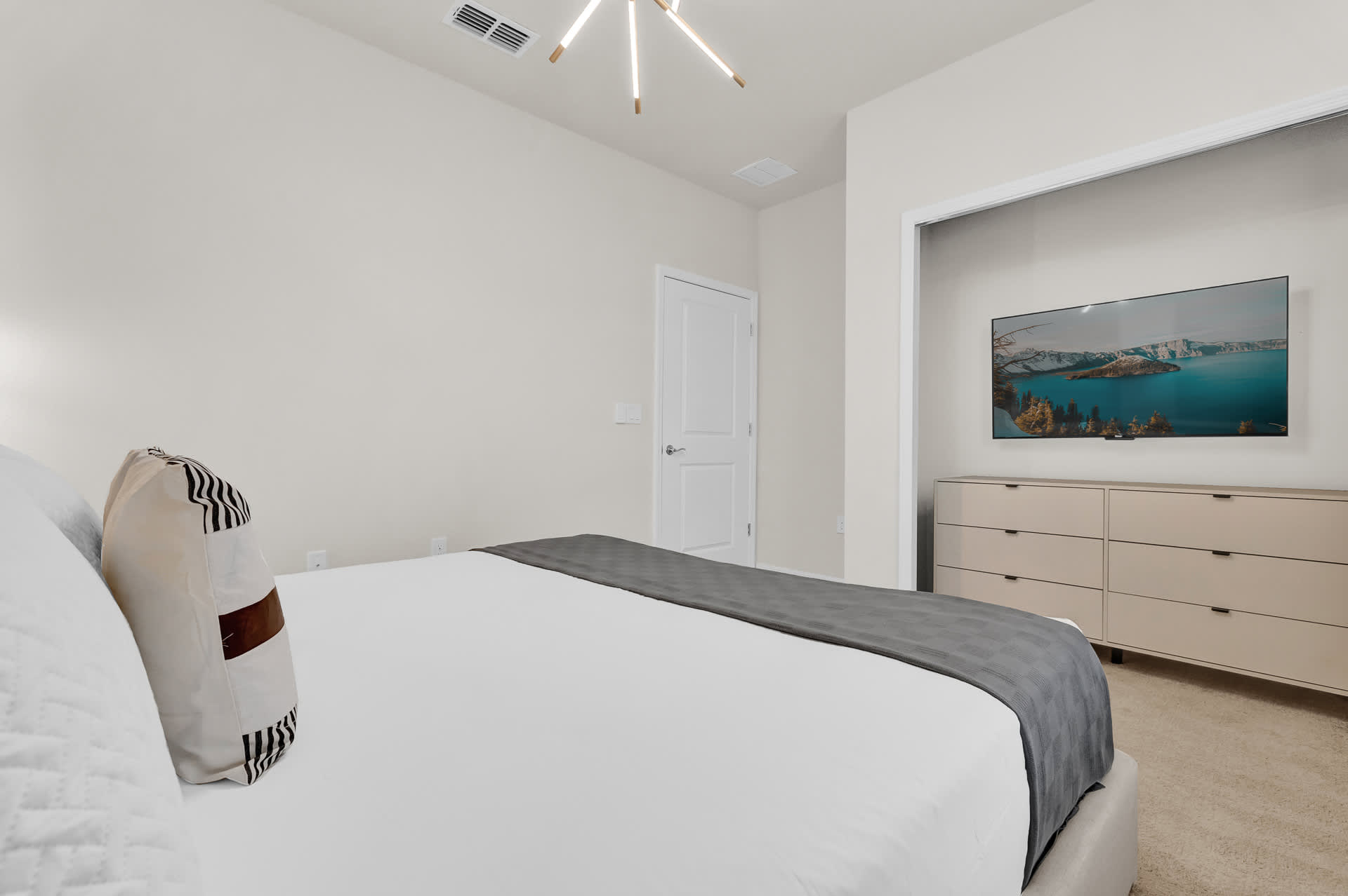 Modern bedroom with light-colored walls and carpet, featuring a wall-mounted TV above a light wood dresser. A white door and a glimpse of the bed with decorative pillows are visible.