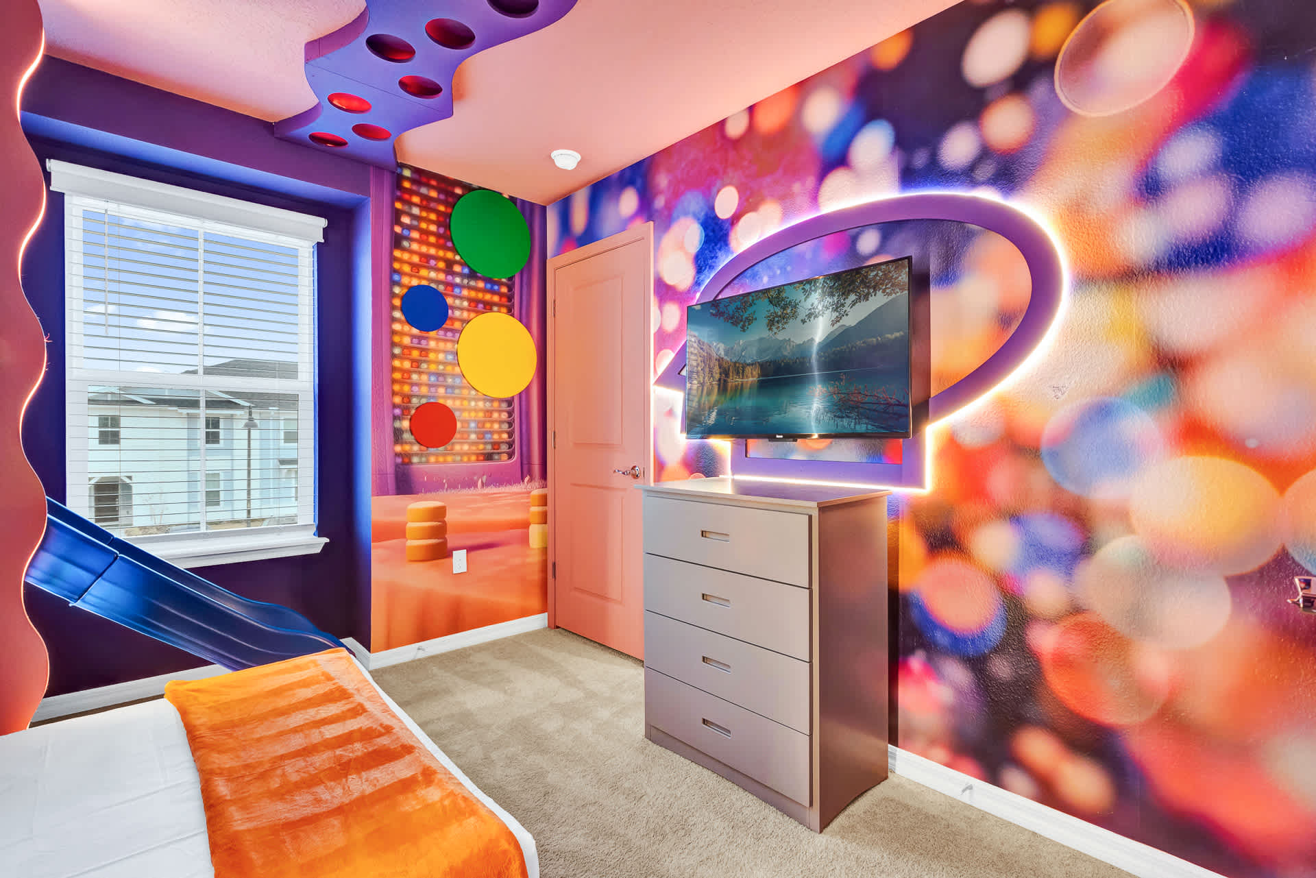 Playful themed bedroom with bright, colorful walls and ceiling, featuring a wall-mounted TV above a dresser. A blue slide and circular wall decor add to the whimsical design.