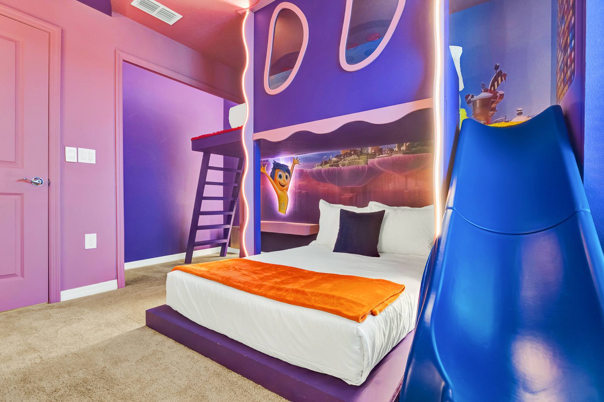 Vibrant themed bedroom with purple walls and ceiling, featuring a low-profile bed, a built-in bunk bed with a ladder, and a blue slide. Character cutouts and colorful lighting create a playful atmosphere.