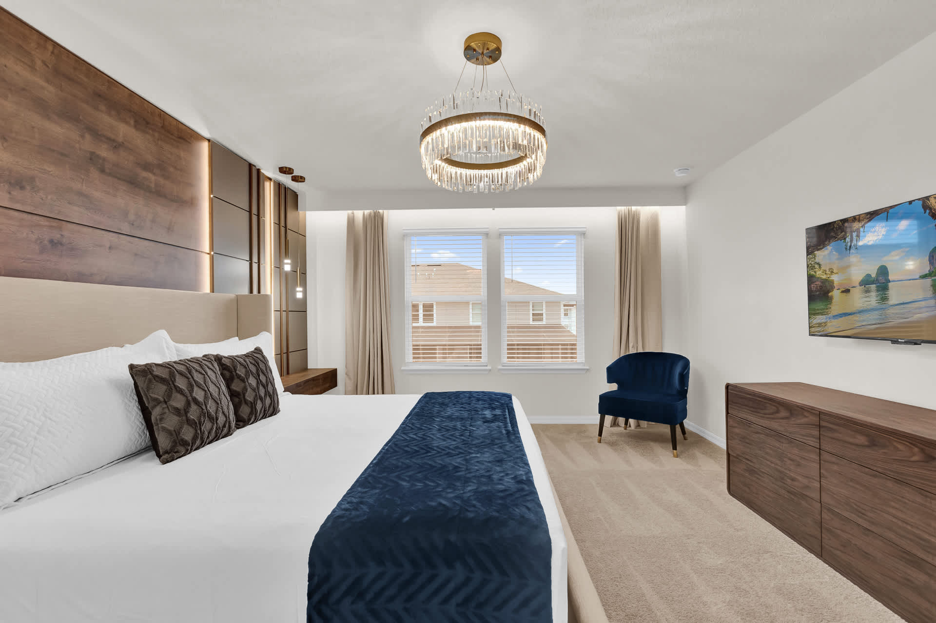 Modern master bedroom featuring a king-sized bed with a beige headboard, dark wood accent wall with integrated lighting, and a stylish gold and crystal chandelier. A blue velvet armchair sits by the window.