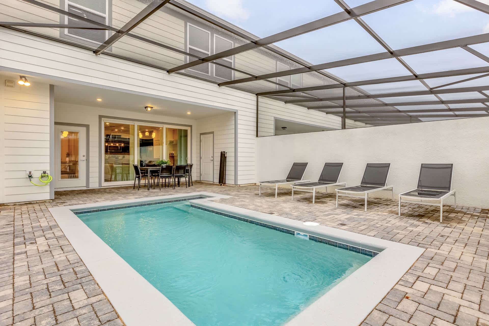 A private outdoor swimming pool is surrounded by light-colored pavers and four lounge chairs, all under a clear, screened enclosure. A covered patio area with an outdoor dining table and chairs is visible next to the house.