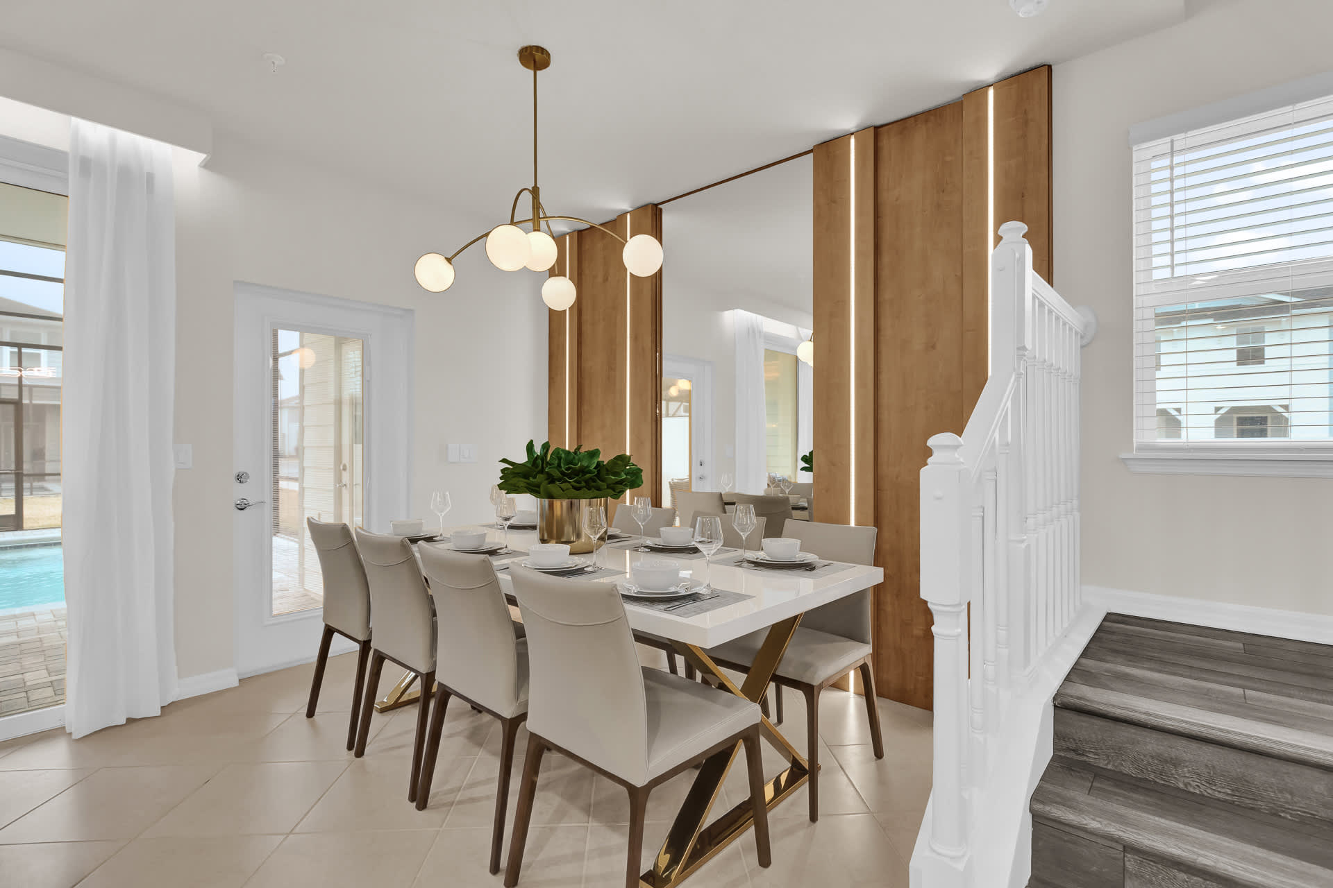 A modern dining room features a long white rectangular table with gold accents, surrounded by eight upholstered dining chairs. A striking gold chandelier hangs above, and a wood-paneled accent wall with integrated mirrors adds depth and style to the space.