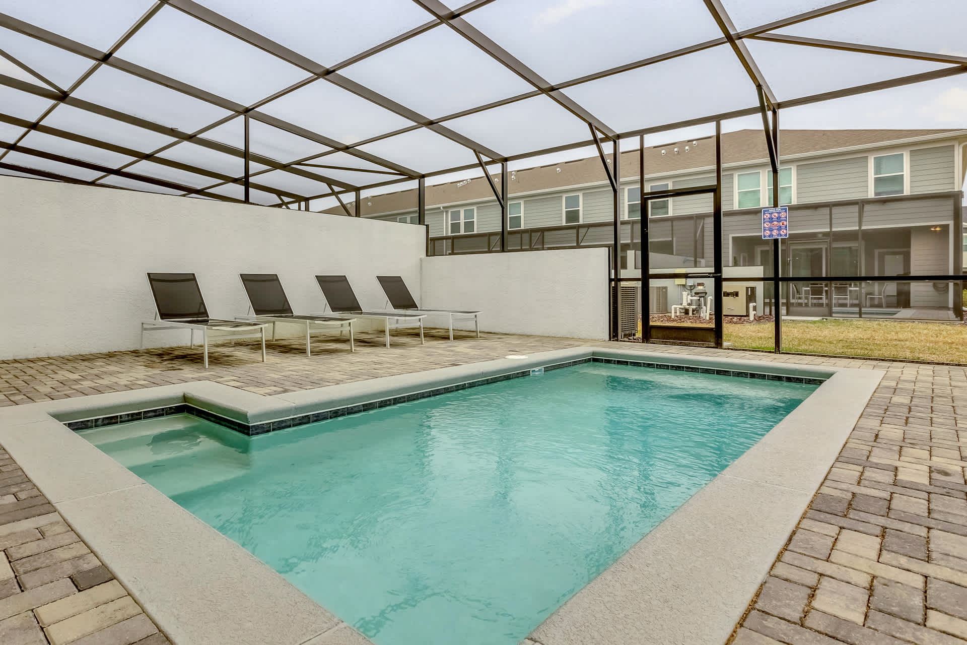 A private, screened-in pool area features a rectangular swimming pool with an attached spa, surrounded by light-colored pavers. Four black lounge chairs are arranged along one side, and a safety fence separates the pool from the adjacent yard.