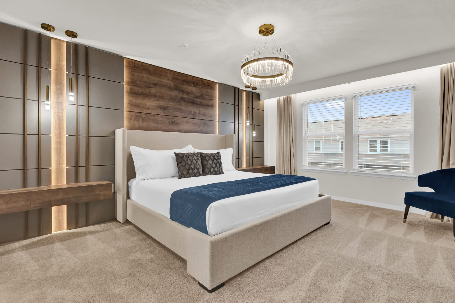 A luxurious bedroom features a king-sized upholstered bed with a blue throw, set against a dramatic dark wood and paneled accent wall with integrated lighting and hanging pendant lights. A modern chandelier illuminates the room, which has carpeted flooring and large windows.