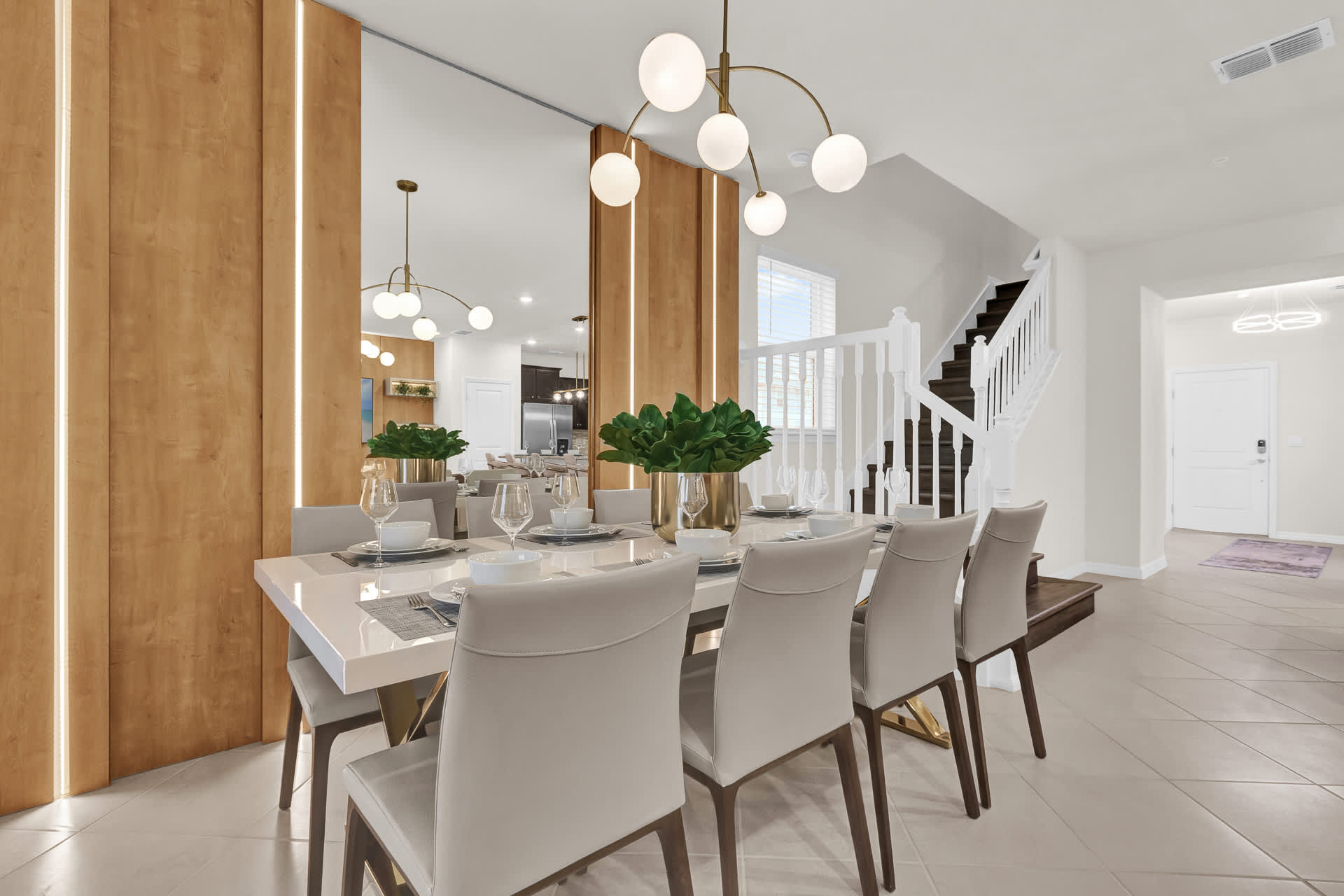 A stylish dining room features a long white dining table set for eight, with upholstered chairs and a modern gold chandelier. A prominent wood-paneled accent wall with integrated mirrors reflects the bright space, and a staircase is visible in the background.