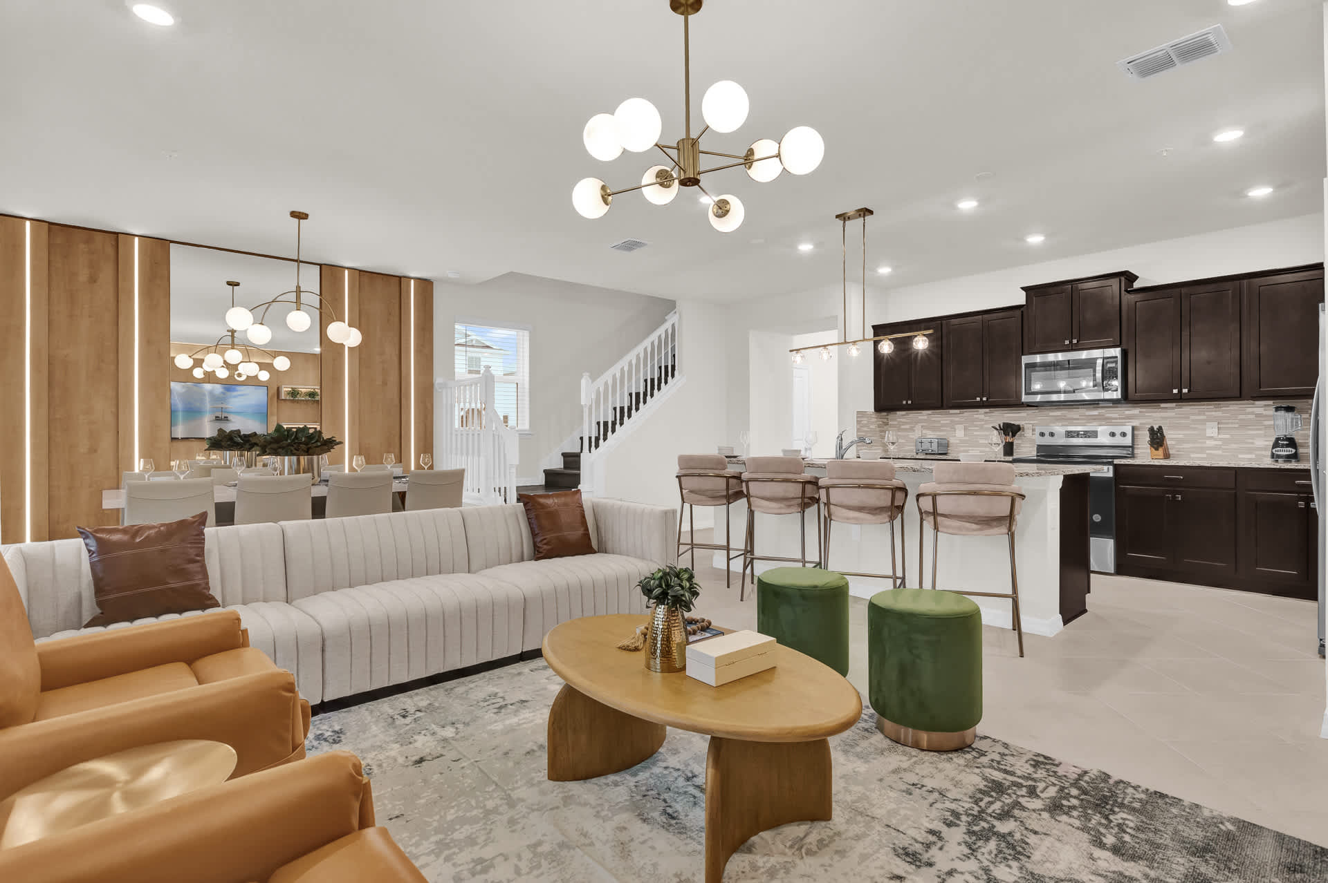 An open-concept living space features a long, light-striped sofa, two tan armchairs, and a modern oval coffee table on a patterned rug. Beyond, a dining area with a large table and a contemporary kitchen with dark cabinetry and a breakfast bar are visible.