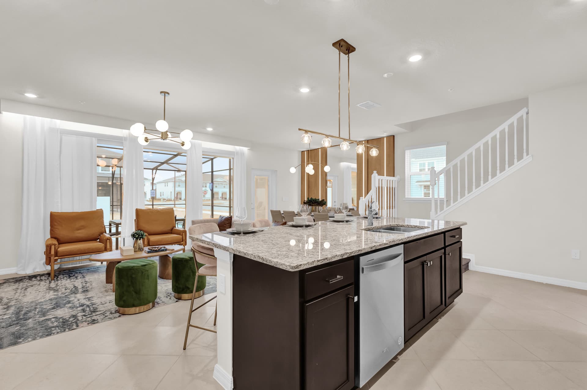 An expansive open-concept living space features a large kitchen island with seating, leading into a bright living room with a wood-paneled accent wall and large windows. A staircase with white railings and dark treads is visible on the right, connecting the levels.