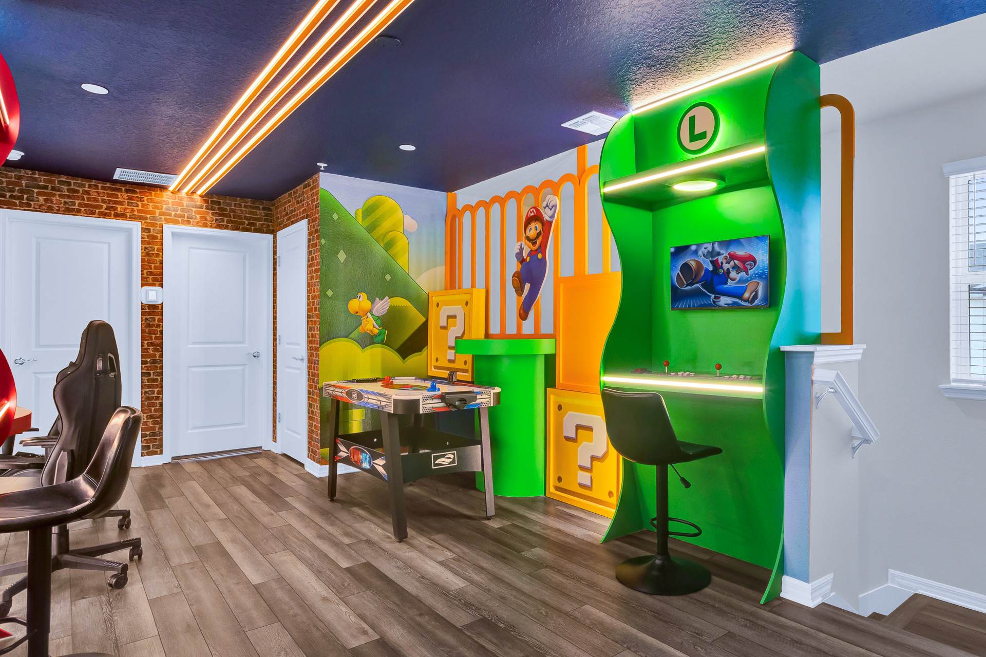 A vibrant, Mario-themed game room features a large green arcade cabinet with a screen, a multi-game table, and two black gaming chairs. The room has dark wood-look flooring, brick accent walls, and bright orange and green wall murals.