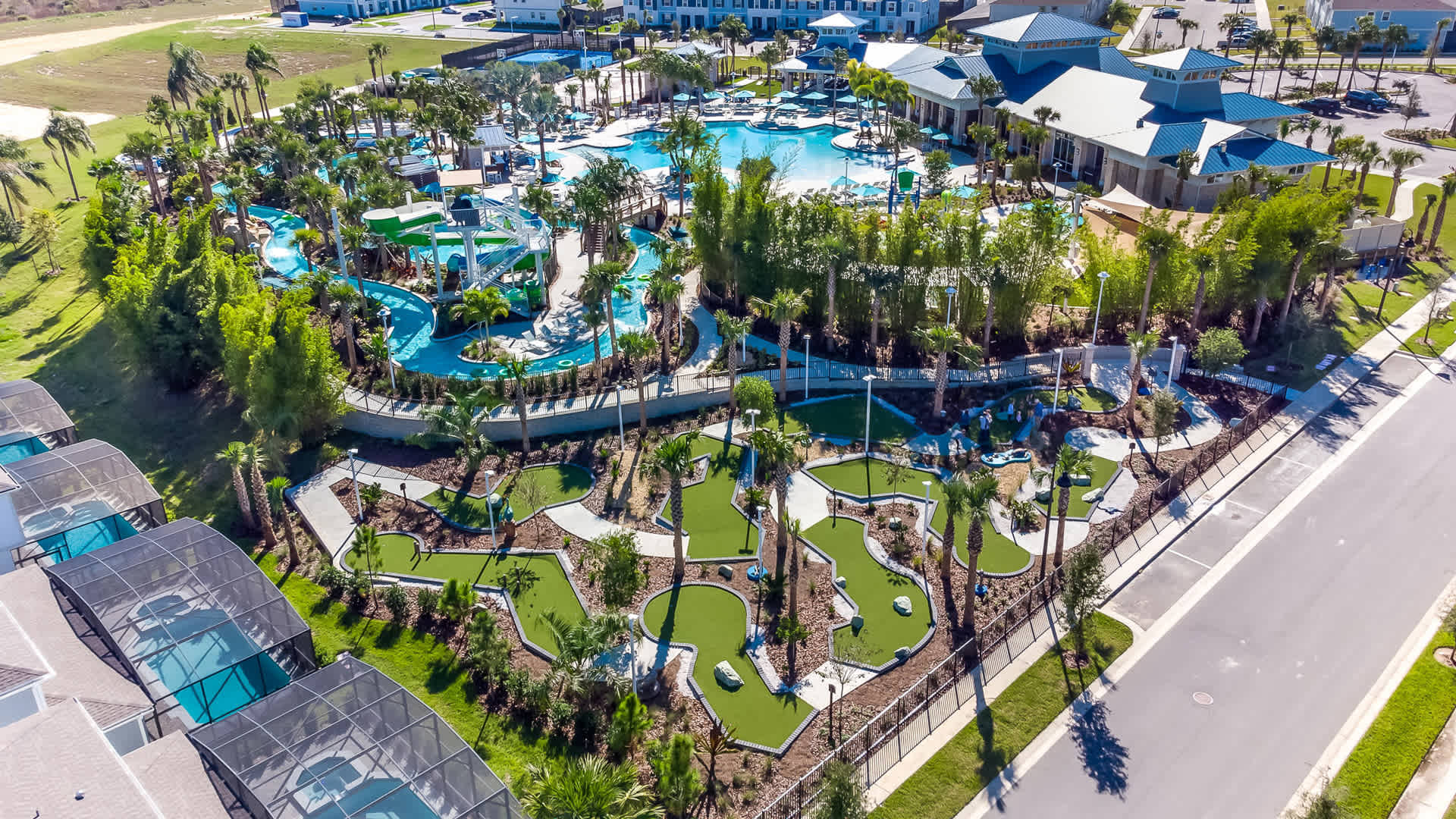 An expansive aerial view showcases a vibrant resort-style water park featuring multiple pools, water slides, a lazy river, and a miniature golf course, surrounded by lush landscaping and residential buildings. The complex offers extensive outdoor recreation.