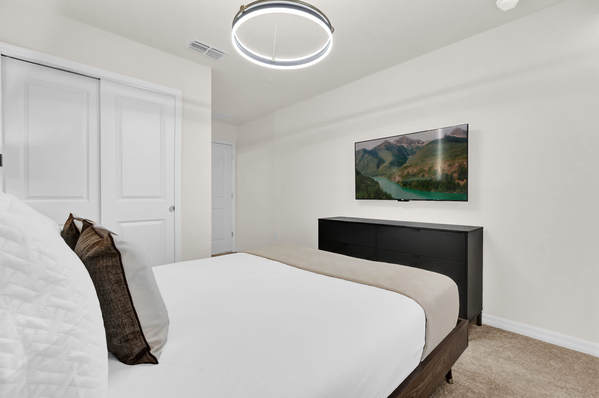 View of a modern bedroom showing a queen bed with white bedding, a dark dresser, and a wall-mounted flat-screen TV displaying a scenic landscape. A large circular chandelier hangs from the ceiling.