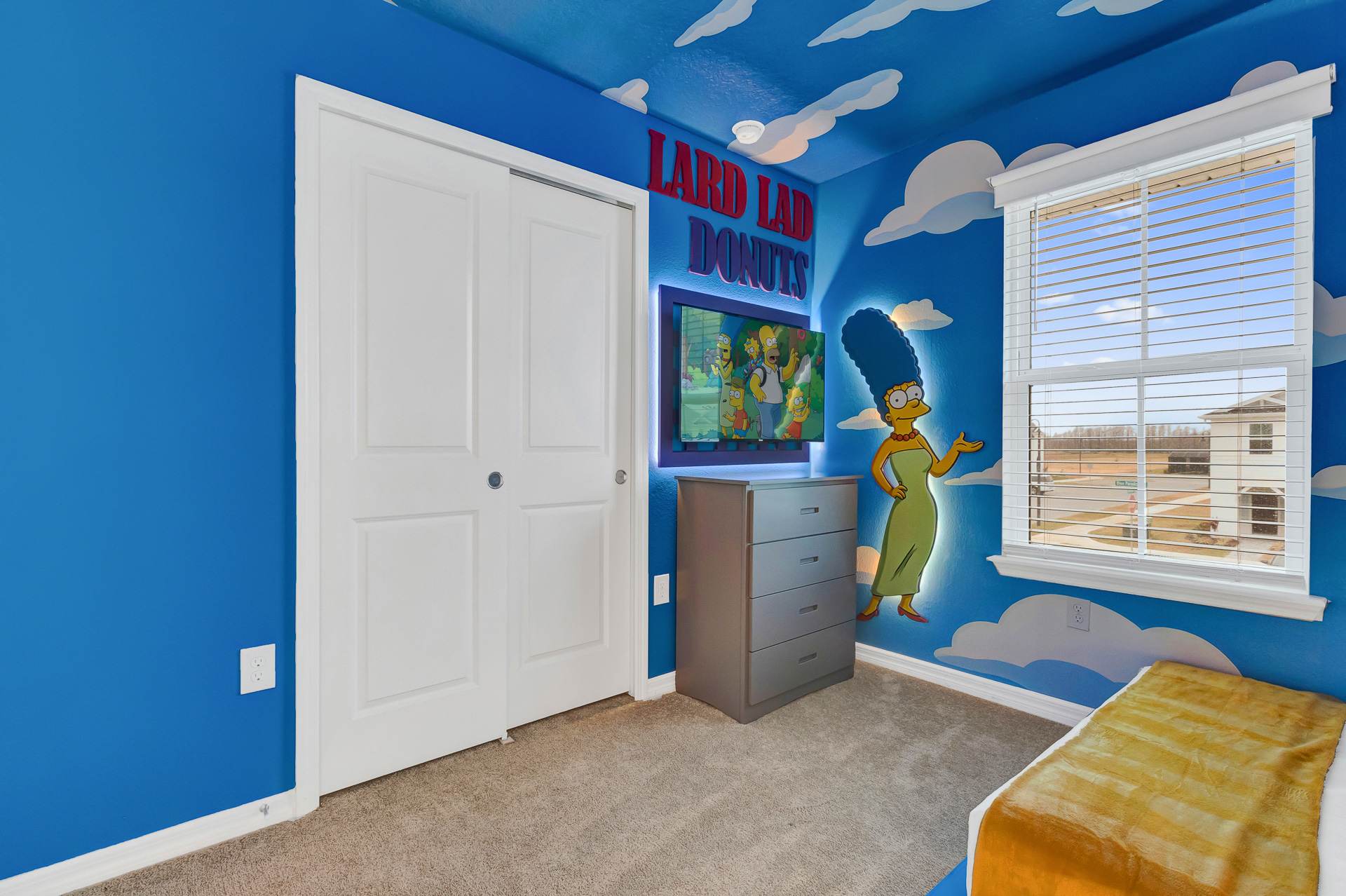 Bright blue themed bedroom with a cloud-painted ceiling, featuring a wall-mounted TV above a gray dresser. A large Marge Simpson cutout and "Lard Lad Donuts" sign decorate the wall next to a white closet.