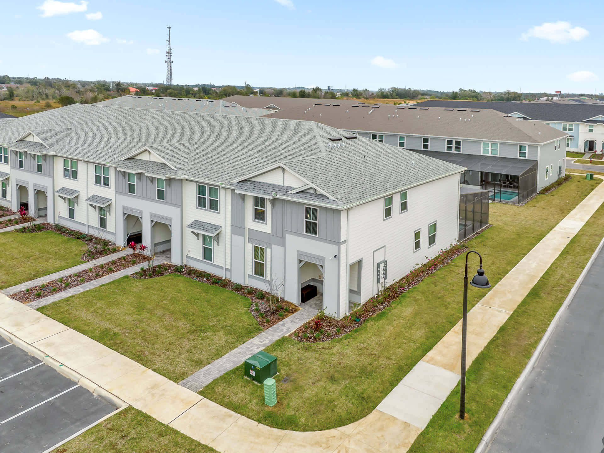 An aerial view shows a row of modern townhouses with light grey and white siding, grey roofs, and small front yards. A paved sidewalk runs along the front, and other similar townhouses are visible in the background.