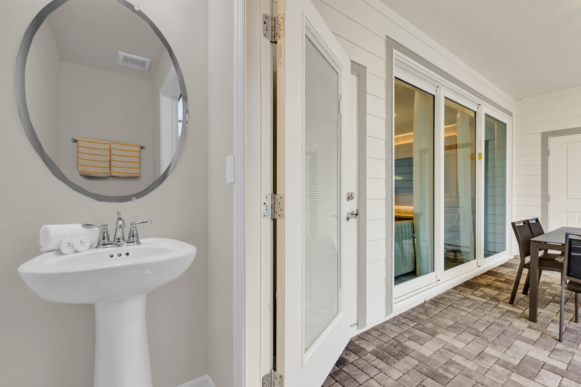 Small bathroom with a white pedestal sink, a round mirror, and yellow striped towels. Adjacent glass doors open to an outdoor patio with gray pavers and a dining set.