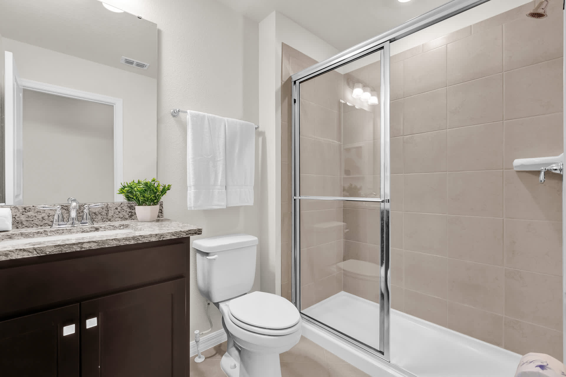 Contemporary bathroom featuring a dark wood vanity with a granite countertop and single sink, a toilet, and a glass-enclosed shower with beige tiled walls. White towels are neatly folded on the vanity.