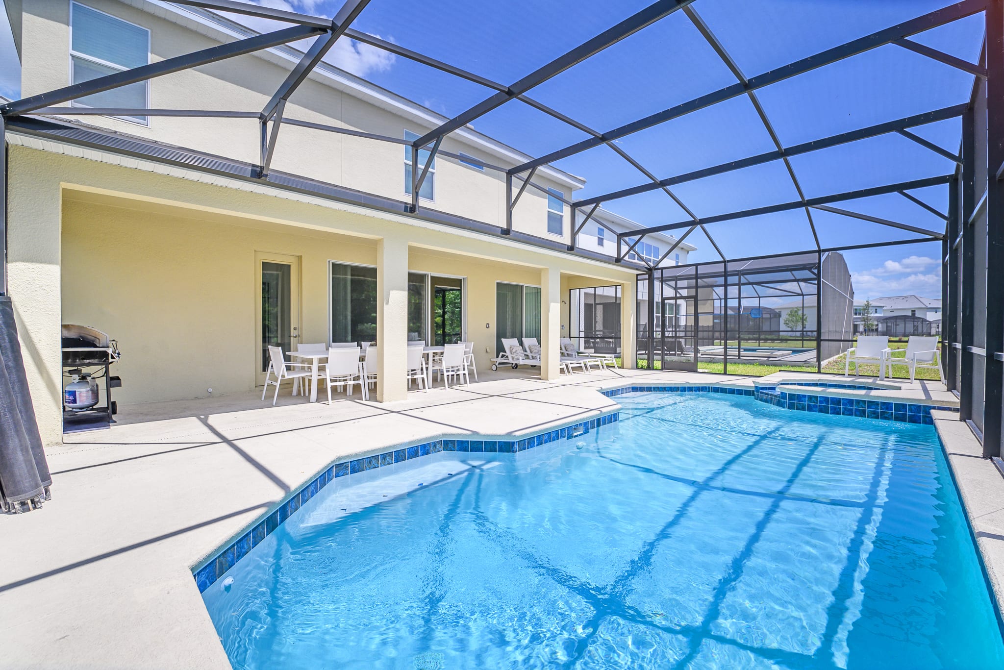 The rear exterior of the house showcases a large screened enclosure covering a private pool and a covered patio area with outdoor dining furniture, all set against a backdrop of lush greenery and a bright blue sky.