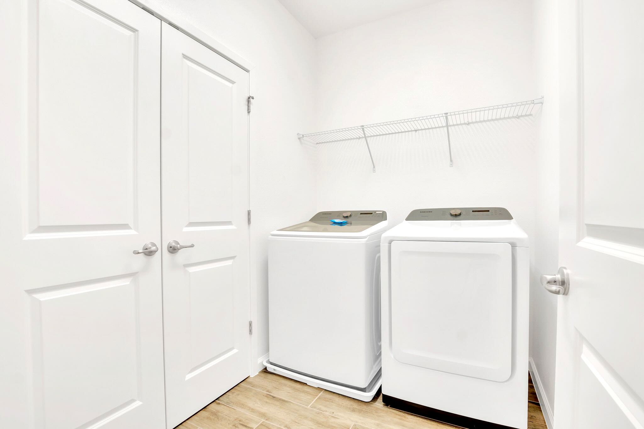 Bright white laundry room with a top-loading washing machine and a front-loading dryer. A wire shelf is mounted above the appliances, and the floor is covered with light wood-look tiles.