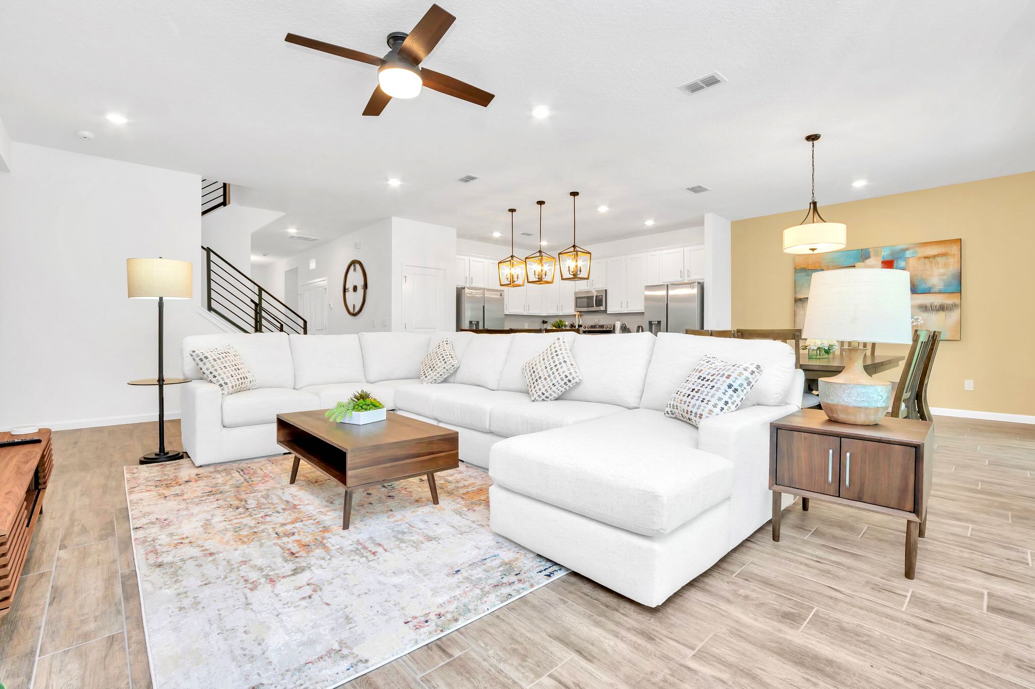A spacious open-concept living room features a large white sectional sofa with patterned throw pillows, a dark wood coffee table on a colorful area rug, and a ceiling fan, with a modern kitchen and dining area visible in the background.