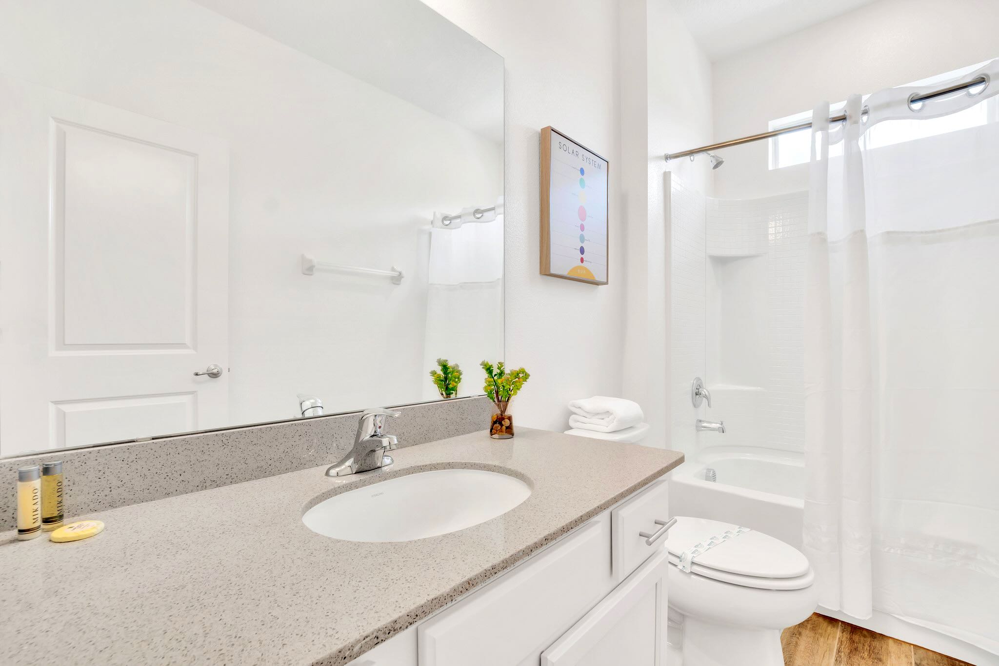 A bright white bathroom with a single vanity, a large mirror, a toilet, and a bathtub with a white shower curtain, accented by a colorful "Solar System" artwork.