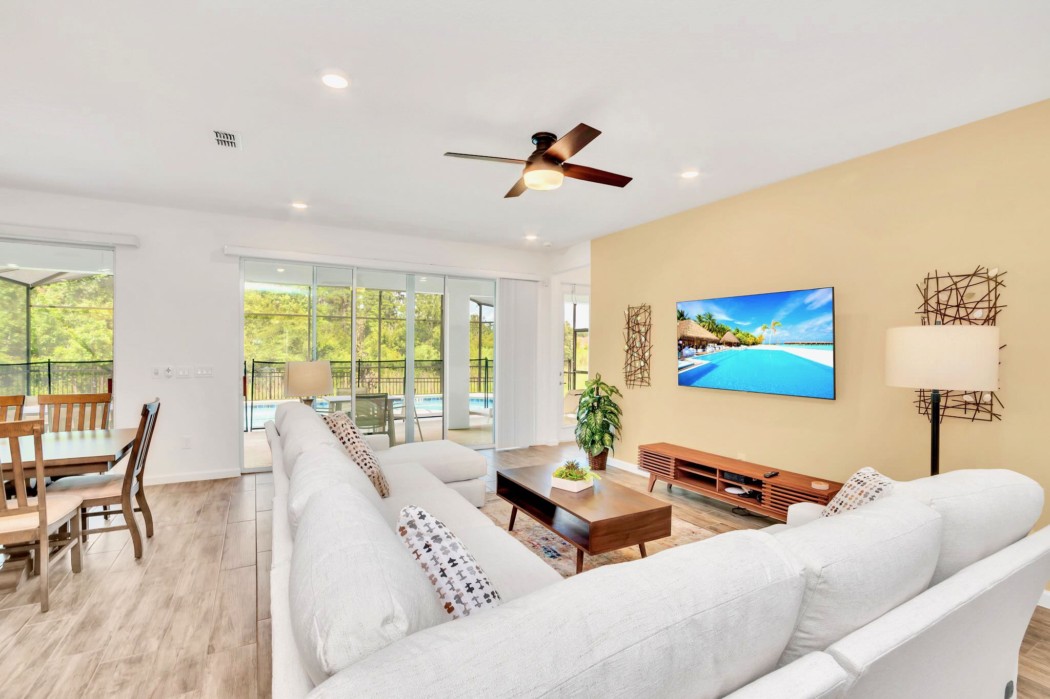 A bright living room with a large white sectional sofa, a wooden coffee table, and a wall-mounted TV, featuring large sliding glass doors that open to a screened-in pool area.