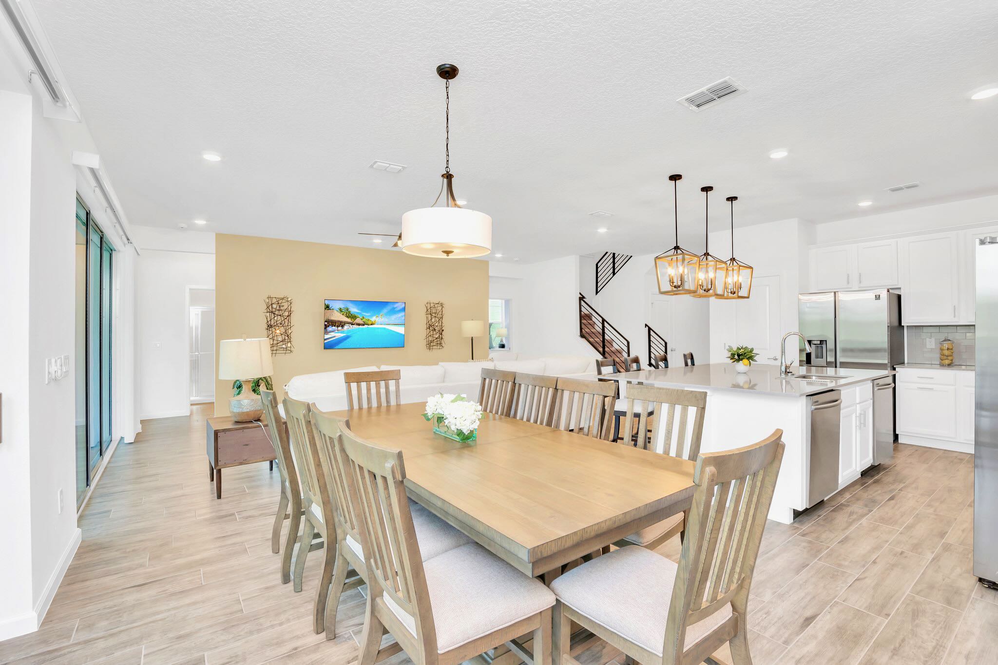 An open-concept dining area with a large wooden table and eight chairs, illuminated by a modern pendant light, adjacent to the living room and kitchen.
