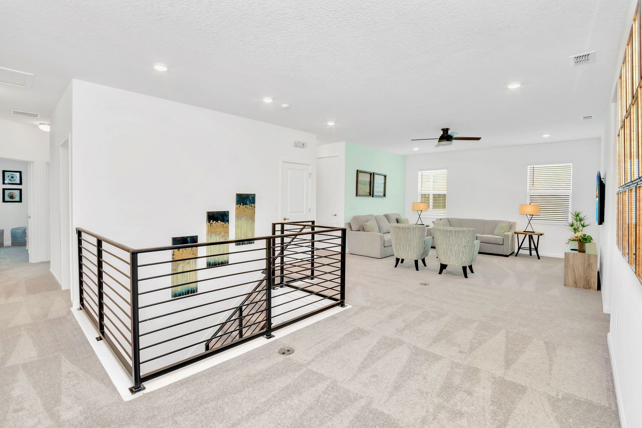 An upstairs loft area with light gray carpet, featuring a modern black metal railing overlooking the stairs, and leading into a secondary living space with seating.