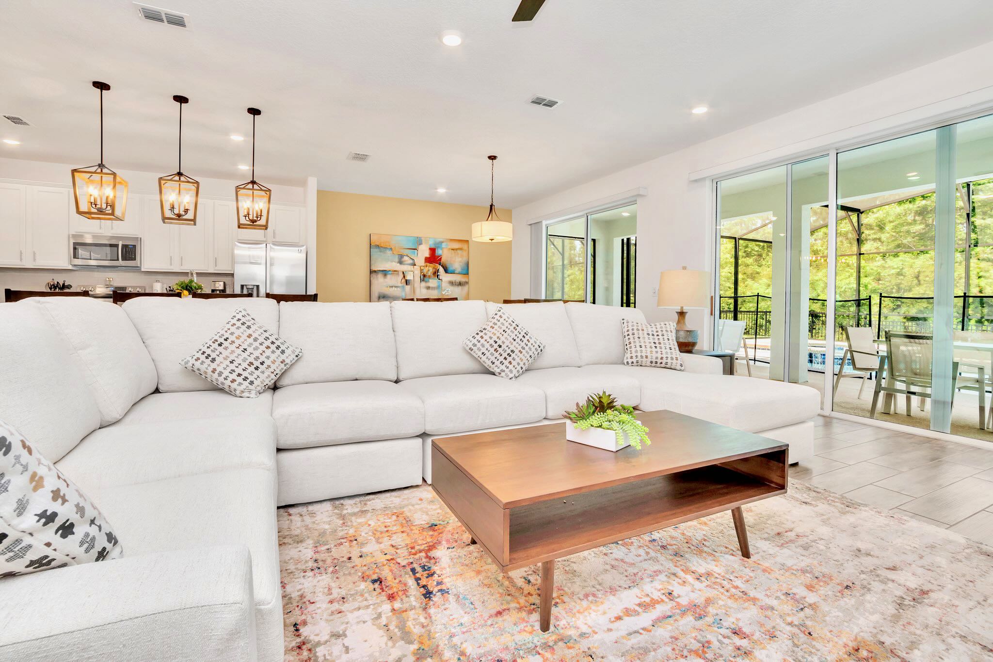 The bright living room features a large white sectional sofa and a wooden coffee table on an area rug, with expansive sliding glass doors opening to the screened patio and pool area, bringing in natural light and outdoor views.