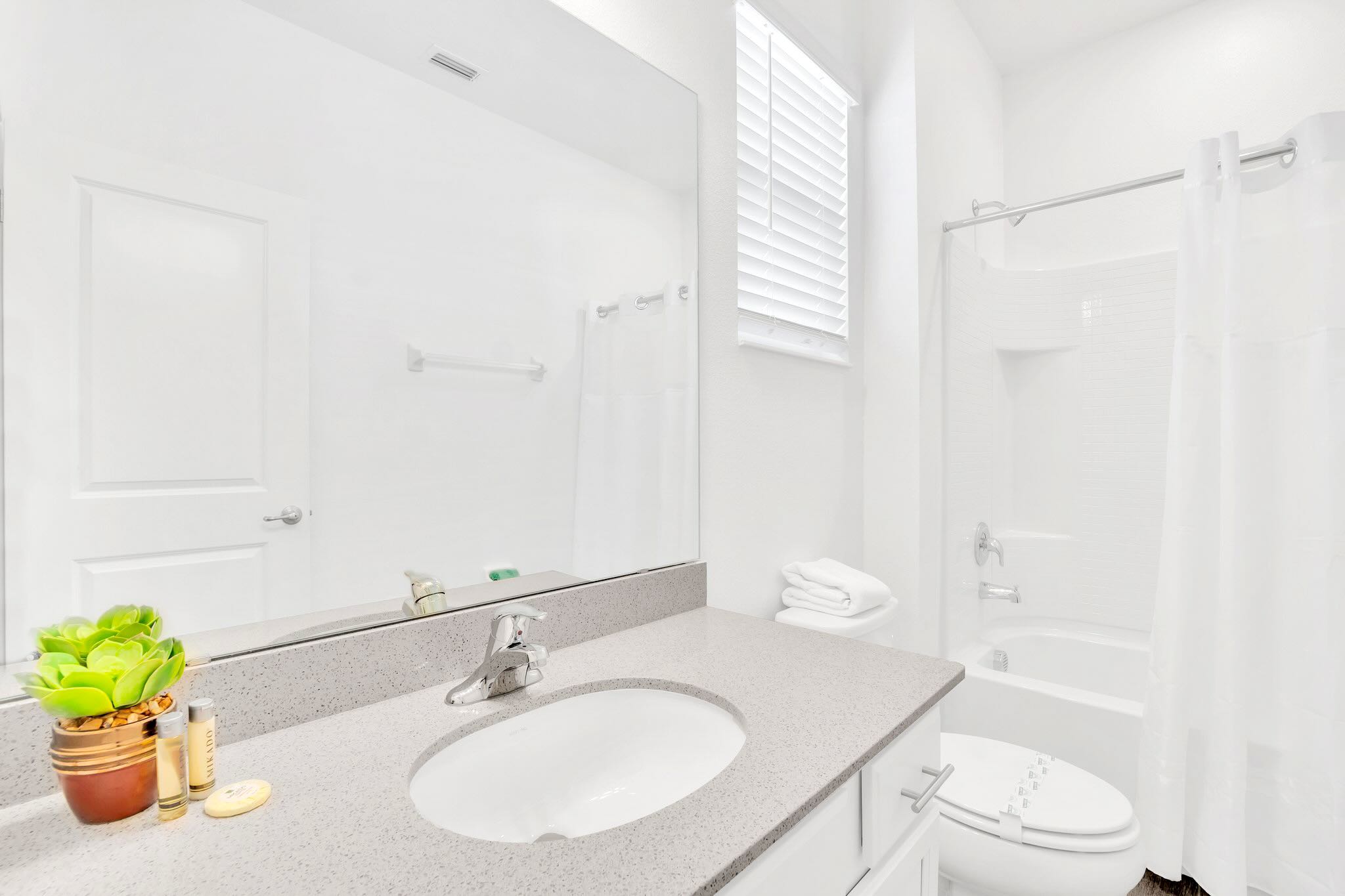 A clean white bathroom with a single vanity featuring a grey speckled countertop, a large mirror, a toilet, and a bathtub with a white shower curtain.