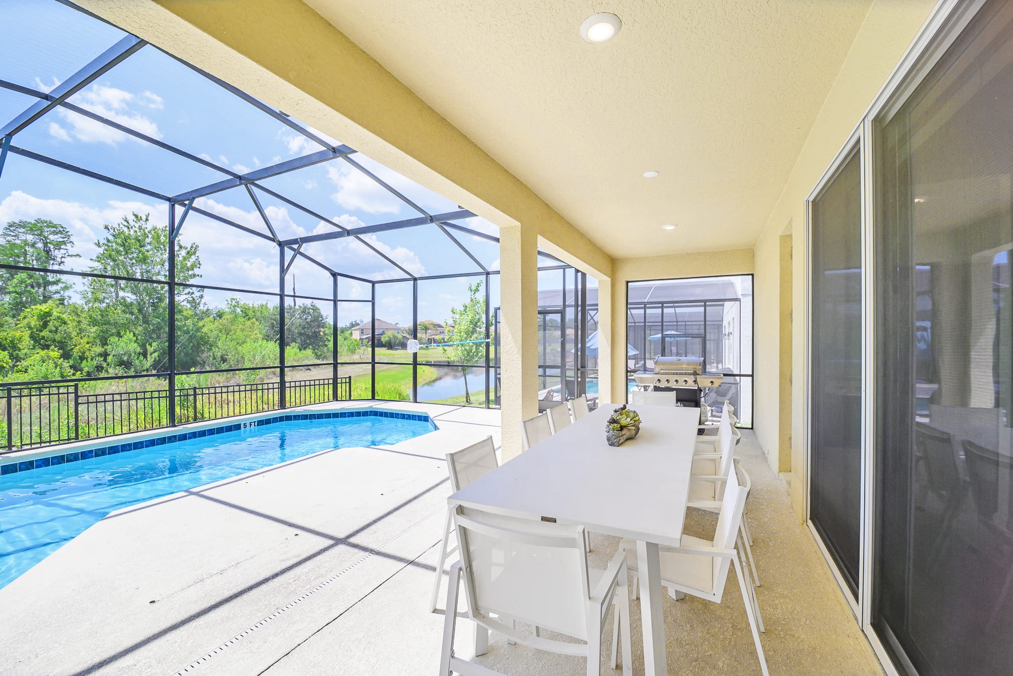 This view of the covered patio highlights a large white outdoor dining table and chairs, a gas grill, and lounge chairs, all within the screened pool enclosure, offering a serene view of the pool and natural landscape.