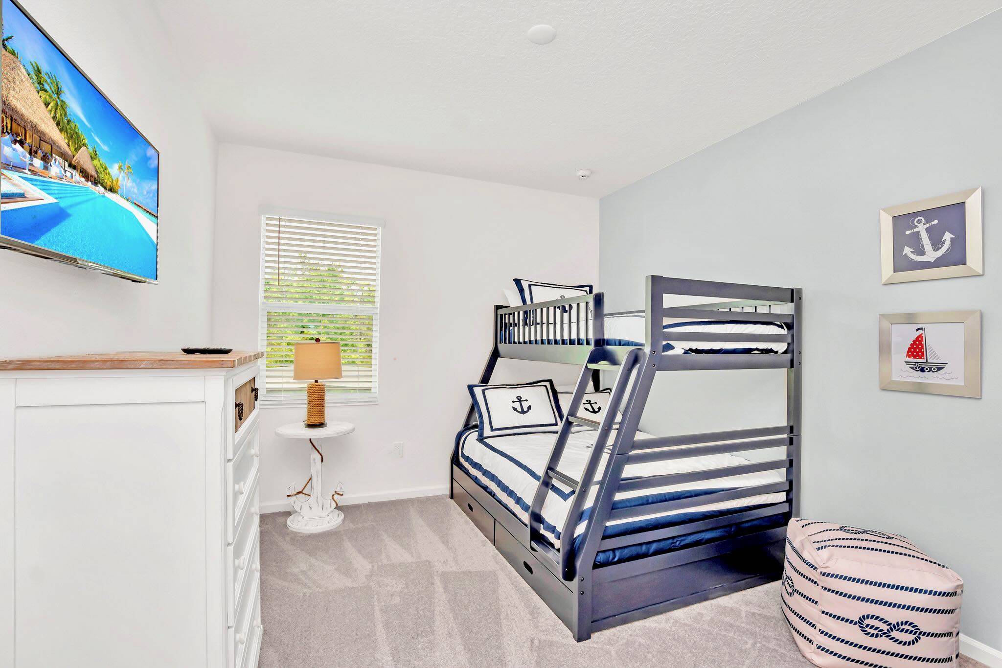 Kids' bedroom with a navy blue bunk bed featuring a nautical theme, a wall-mounted TV, and a white dresser. The room has light gray carpet, a window with blinds, and a light blue accent wall.
