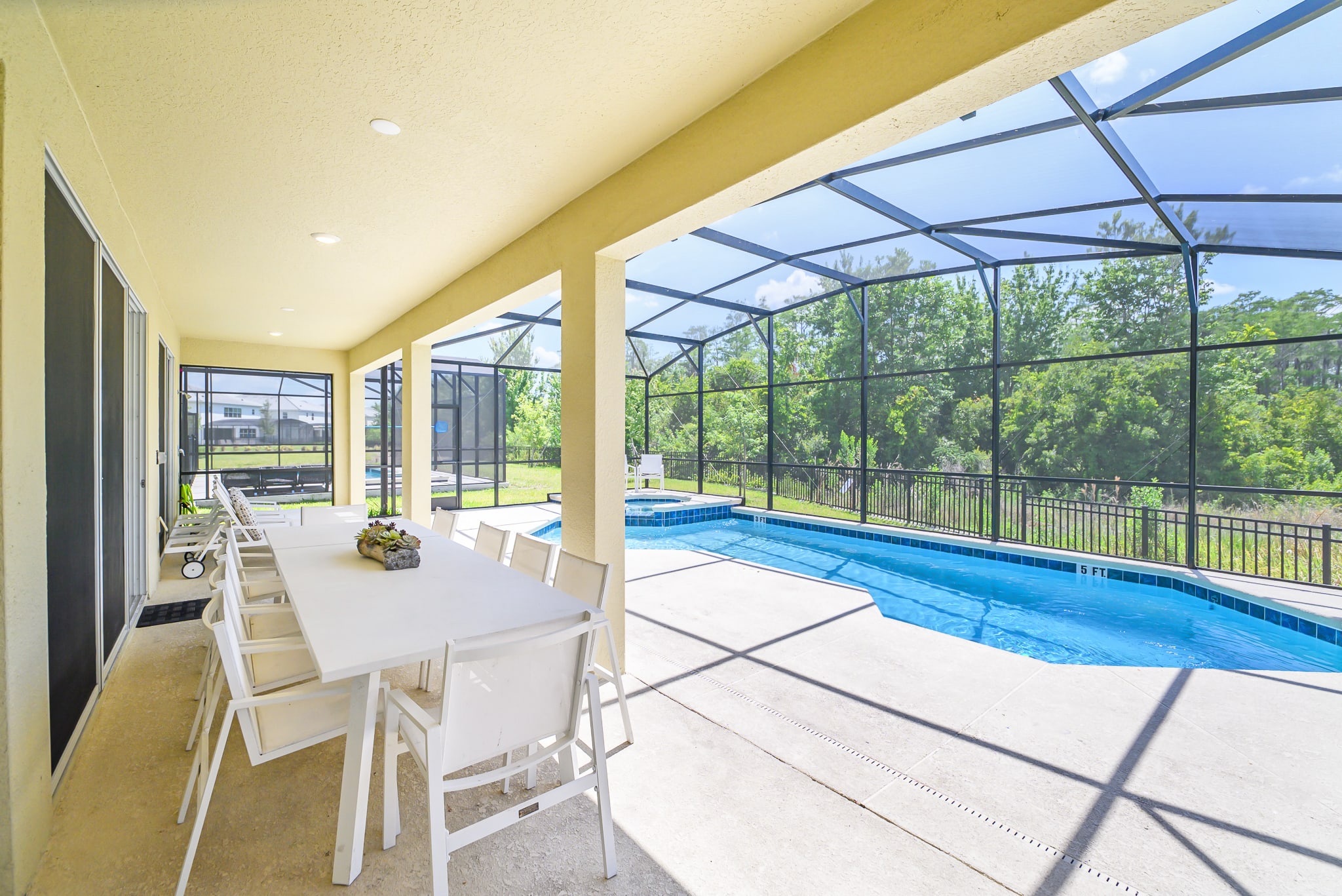 A spacious covered patio features a long white outdoor dining table with multiple chairs, offering a shaded area next to the screened private pool, with views of a tranquil lake and distant trees.