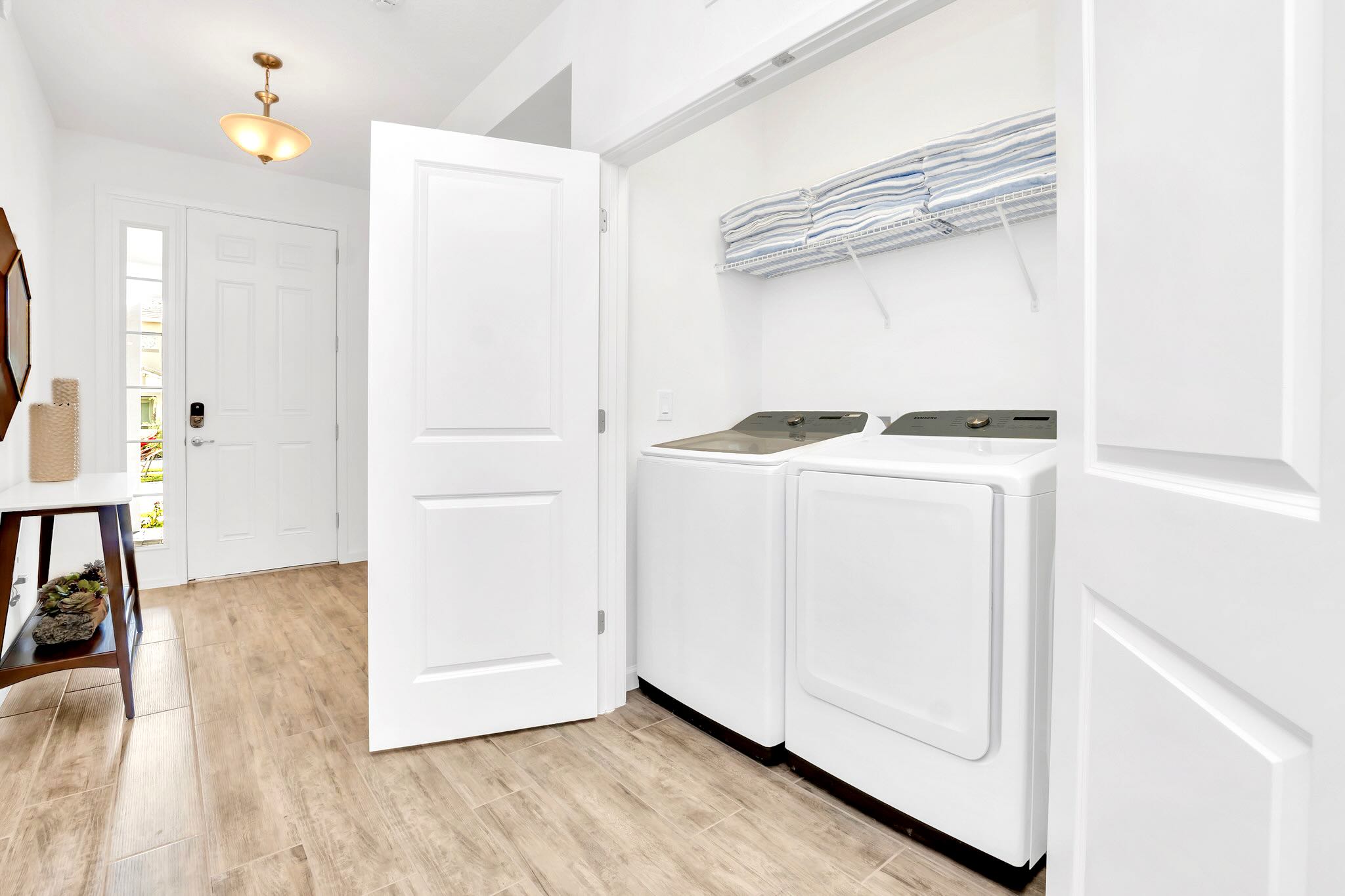 Clean laundry area with a white washing machine and dryer, featuring a wire shelf stocked with neatly folded towels. The space has light wood-look tiled flooring and white doors, with a glimpse of the entryway.